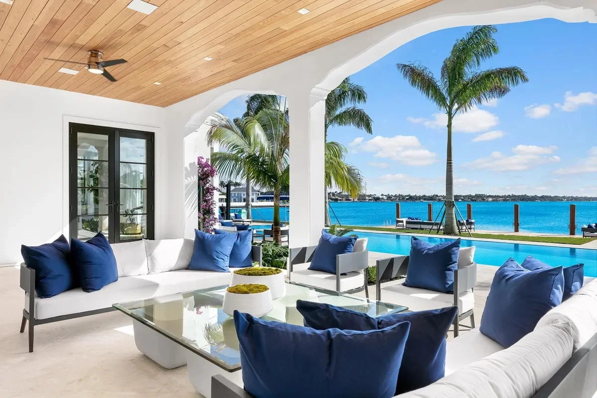 Luxurious patio overlooking a turquoise pool and ocean, with white sofas and blue cushions. Palm trees and clear skies create a serene, tropical vibe.