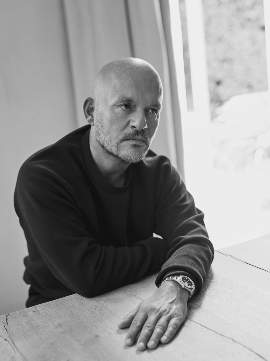 Vincent Van Duysen sits by a wooden table in a bright room, wearing a dark sweater and watch. The black and white image conveys a reflective mood.