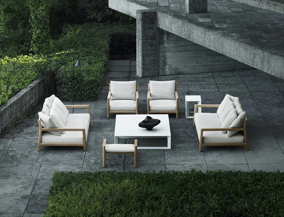 Minimalist outdoor seating area with white cushioned chairs and a sofa around a white coffee table on a stone patio, surrounded by lush greenery.
