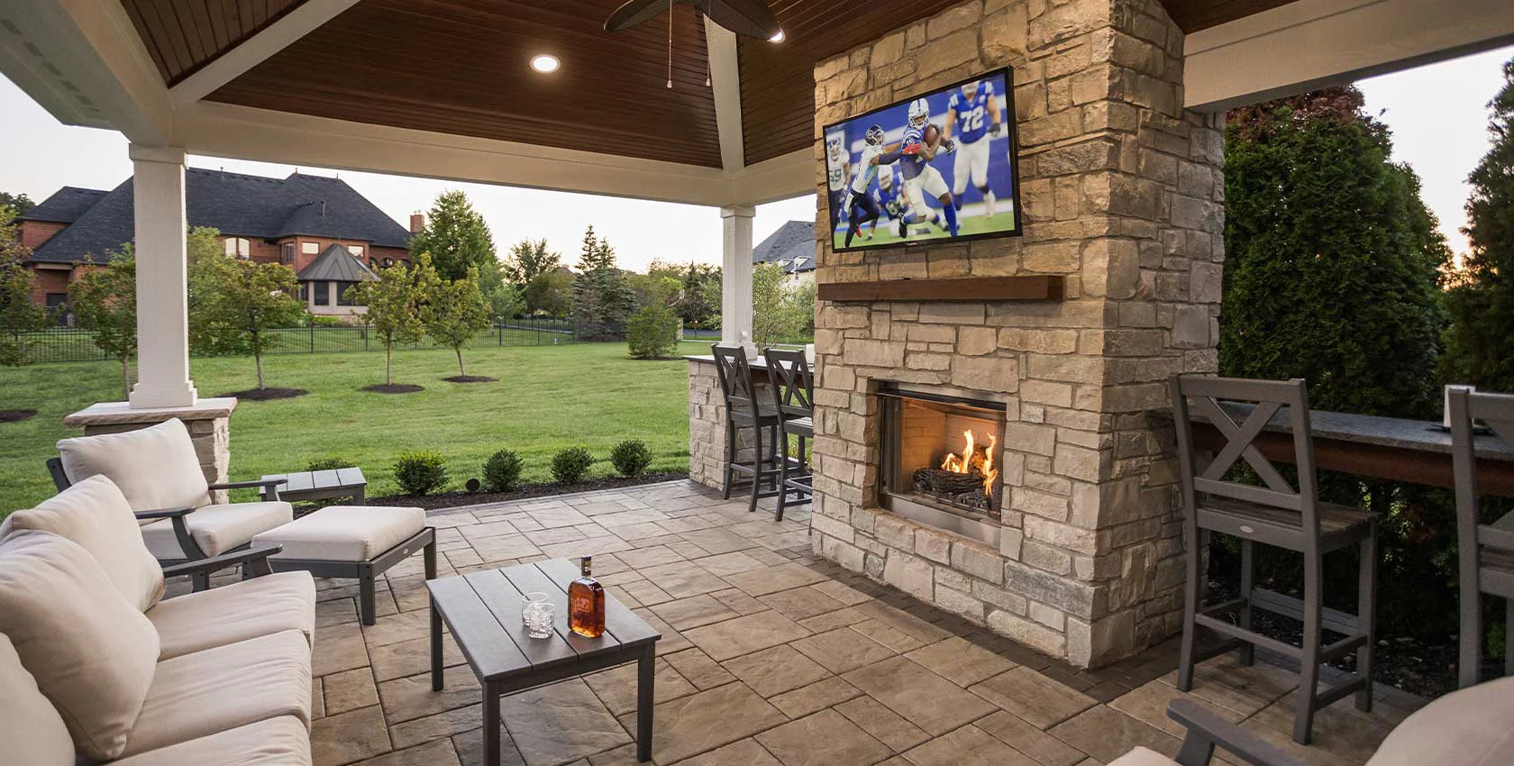 Covered patio with outdoor fireplace and TV. Cozy seating area with light cushions faces the fireplace. Lush green lawn and trees in the background.