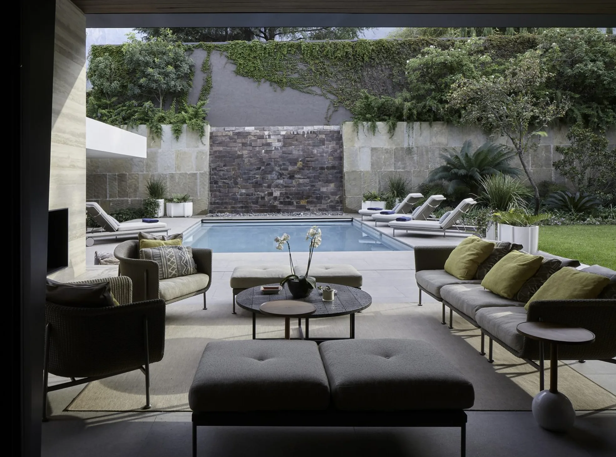 Modern patio with plush seating overlooks a serene pool, surrounded by greenery and stone walls, creating a tranquil and inviting atmosphere.