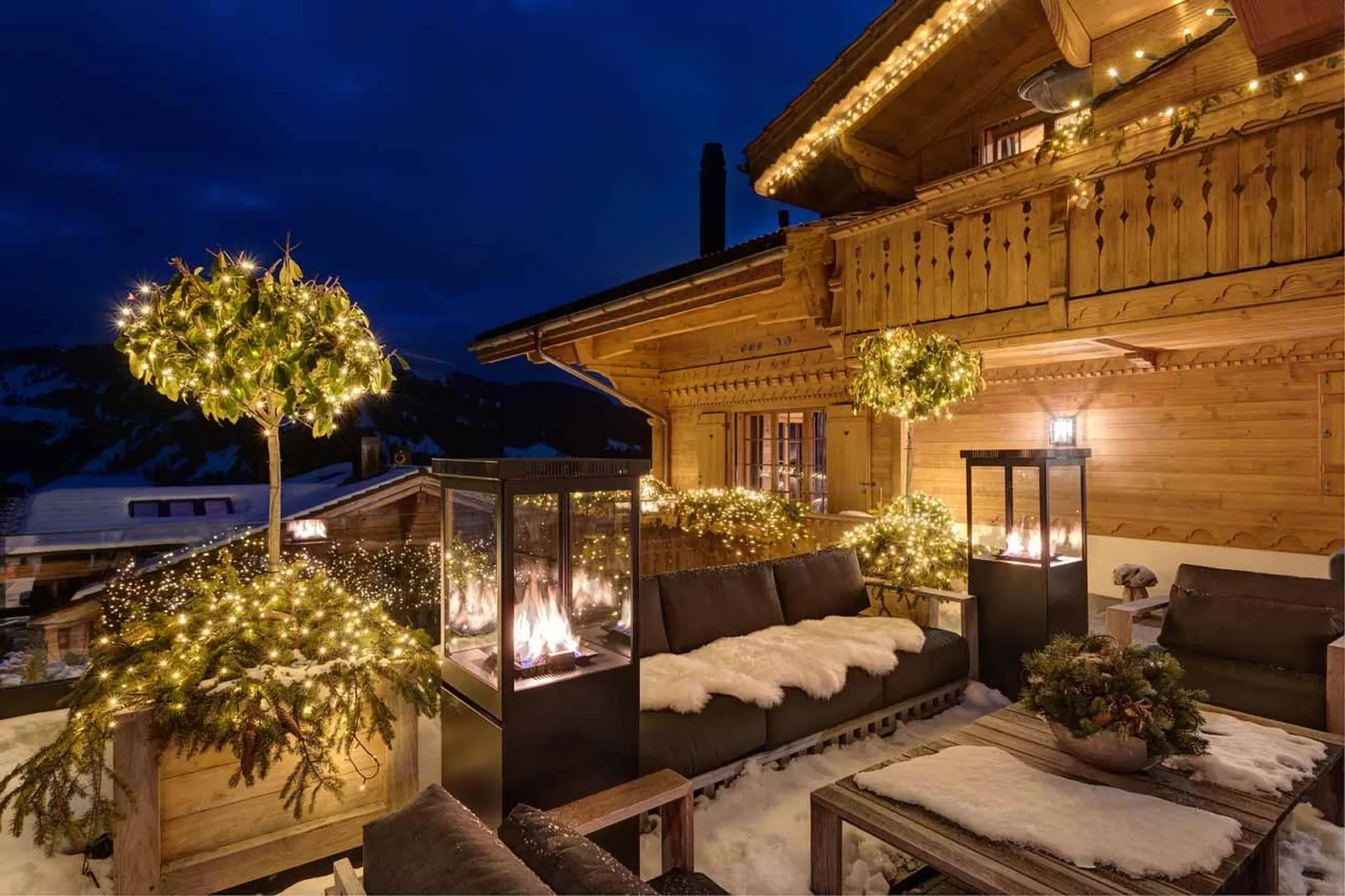 Luxury destionations: cozy outdoor chalet deck at night with warm string lights, soft seating, fur blankets, and glowing fire pits, surrounded by snowy mountain views.