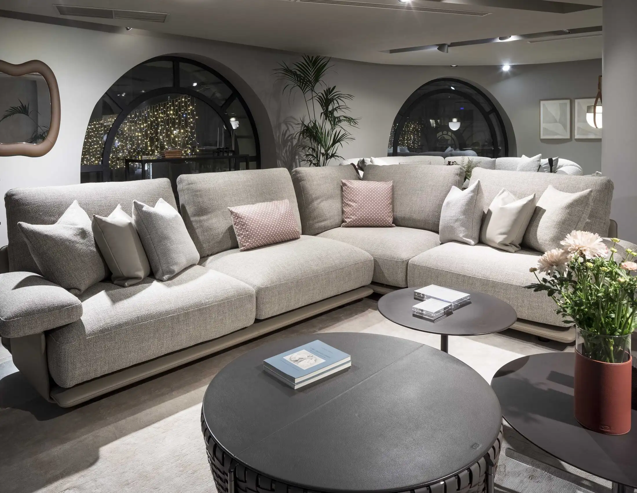 A cozy living room with a large gray sectional sofa adorned with pink and gray cushions. Two round coffee tables hold books and flowers, with warm lighting and decorative arched windows in the background.
