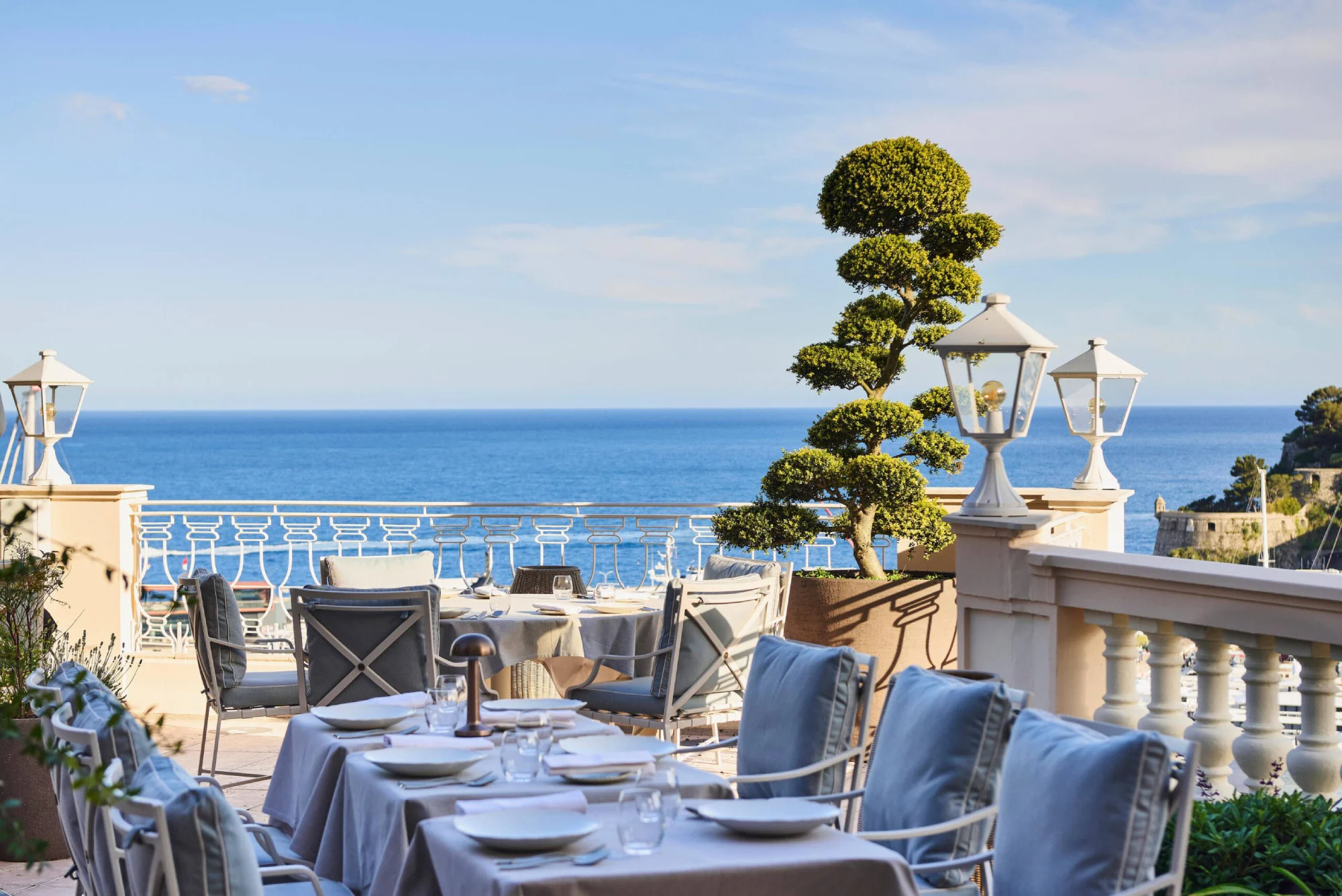 Ocean-view terrace with elegant dining tables set for a meal. Cushioned chairs, topiary plants, and white lanterns create a serene, luxurious atmosphere, typical of luxury hotels.