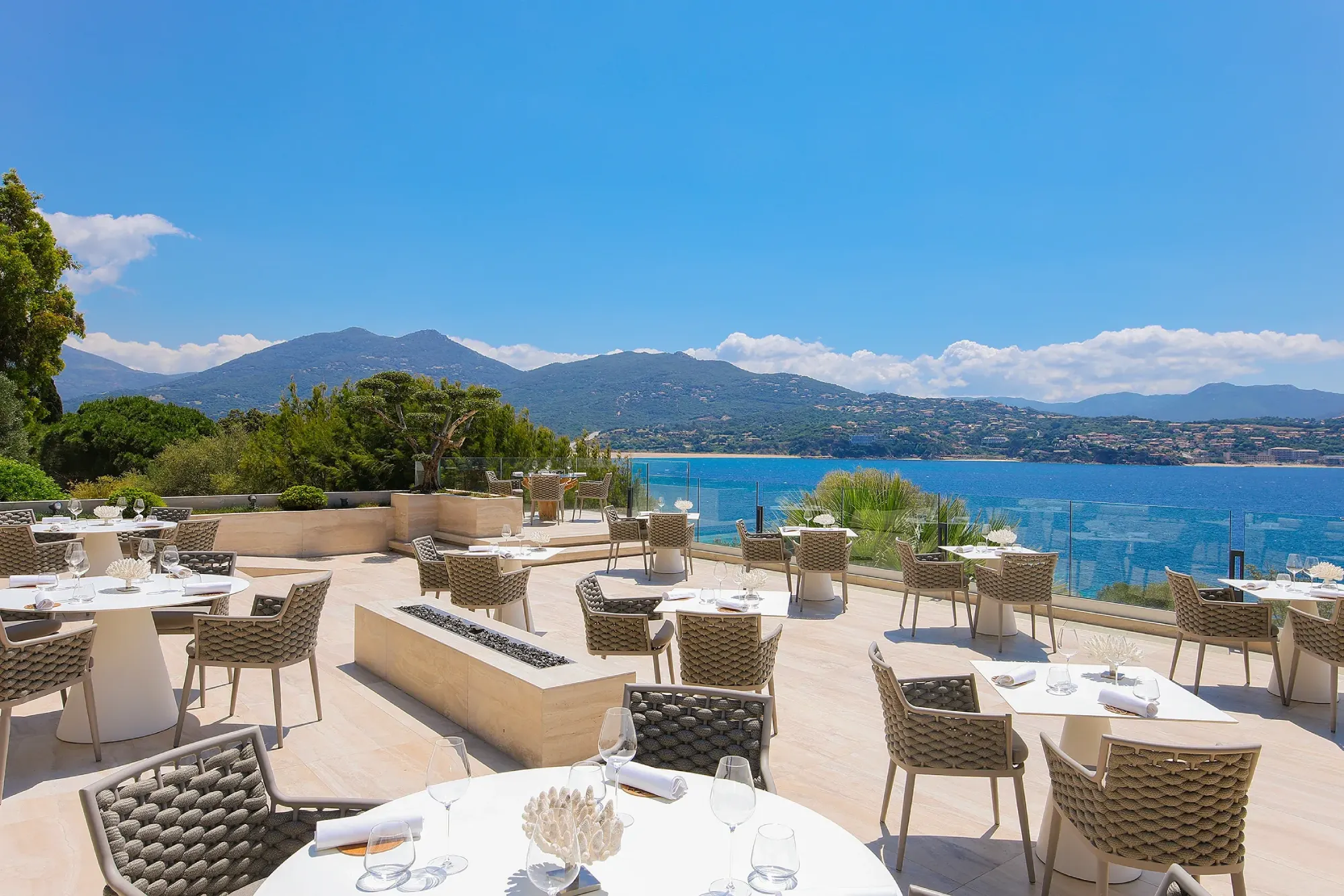 Outdoor restaurant terrace overlooking a serene blue sea and distant mountains under a clear sky. Tables set elegantly, conveying a tranquil ambiance.