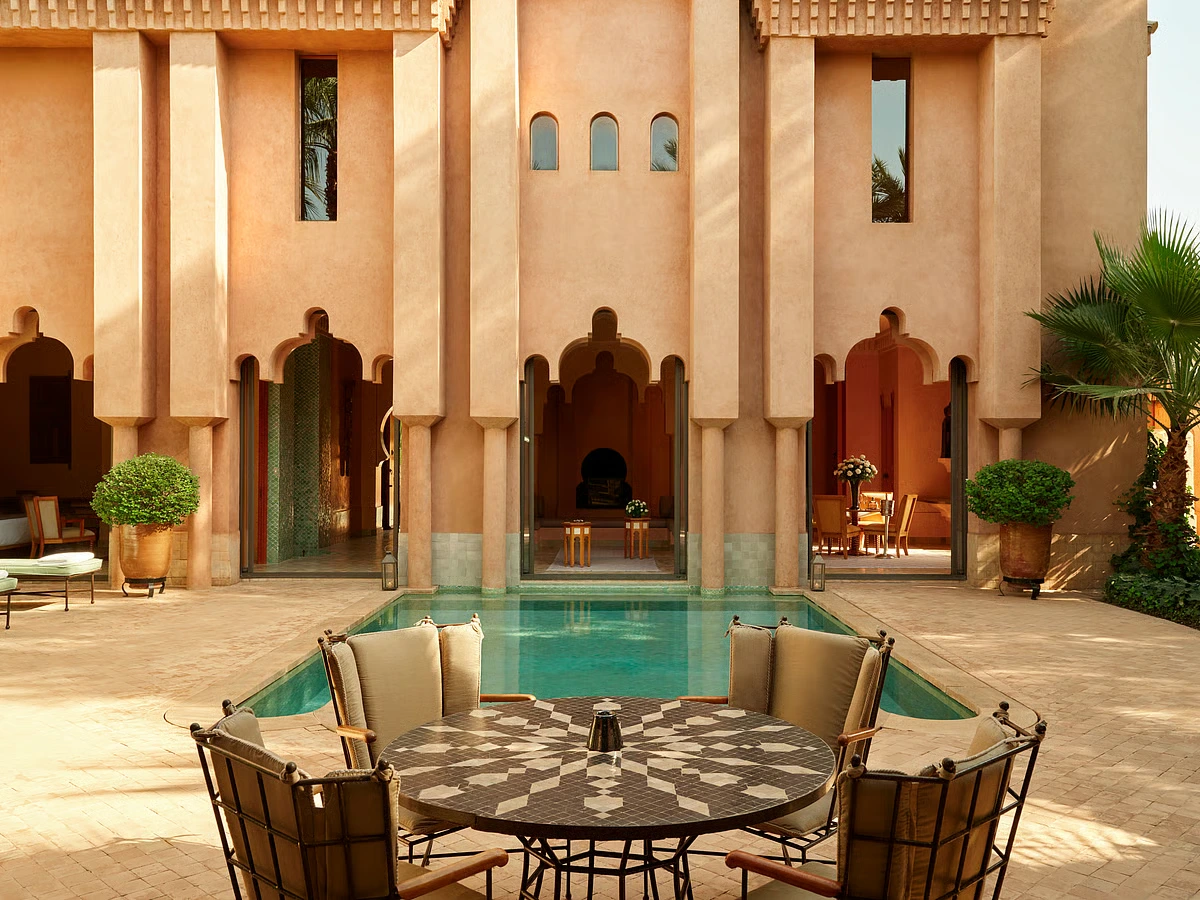 Elegant Moroccan courtyard with arched windows, a central pool, and a mosaic table surrounded by cushioned chairs. Warm, inviting atmosphere.