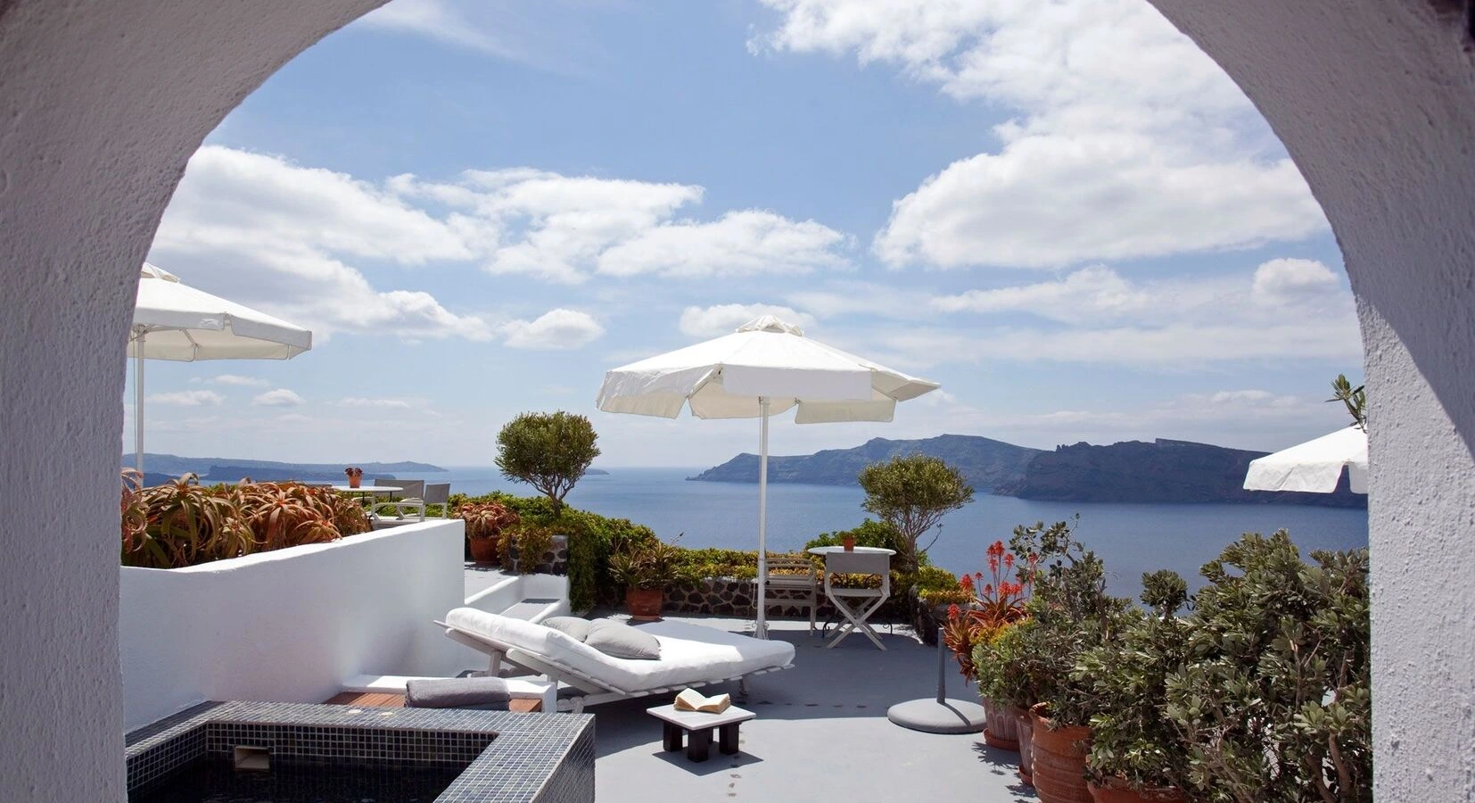Scenic terrace overlooking a serene blue sea and distant islands. White lounge chairs, umbrellas, and lush plants create a tranquil and relaxing atmosphere.