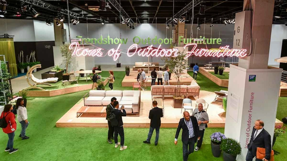Attendees explore a vibrant exhibition of outdoor furniture with green grass flooring, modern seating, and a neon sign reading "Trendshow Outdoor Furniture."