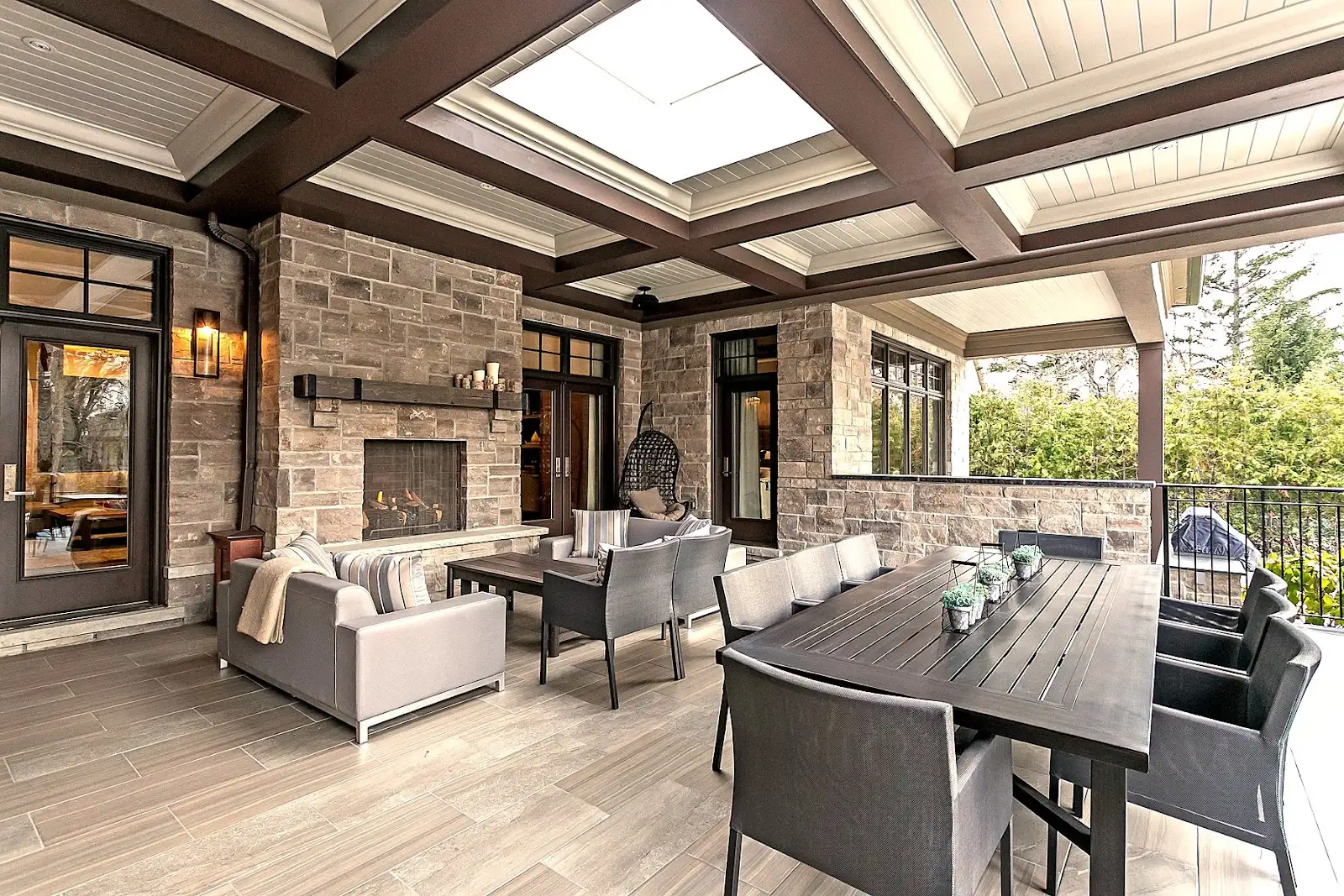 Elegant outdoor patio featuring a stone fireplace, cozy seating area with sofas, wooden dining table with chairs, and a covered ceiling with beams.