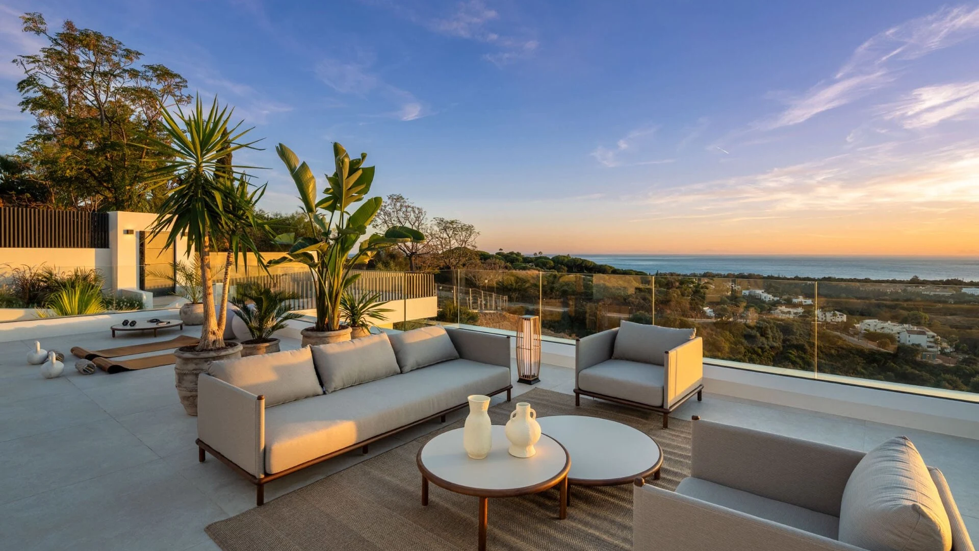Modern rooftop patio with stylish gray furniture, tropical plants, and a stunning ocean view at sunset. The scene conveys tranquility and luxury.