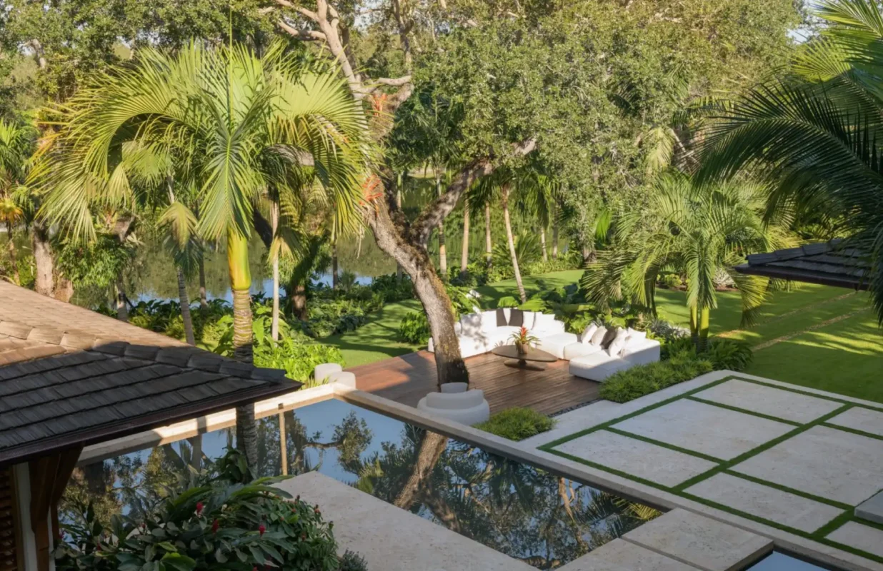 Lush garden with palm trees surrounds a serene patio featuring white cushioned seating and a reflective pool, creating a tranquil, tropical ambiance.