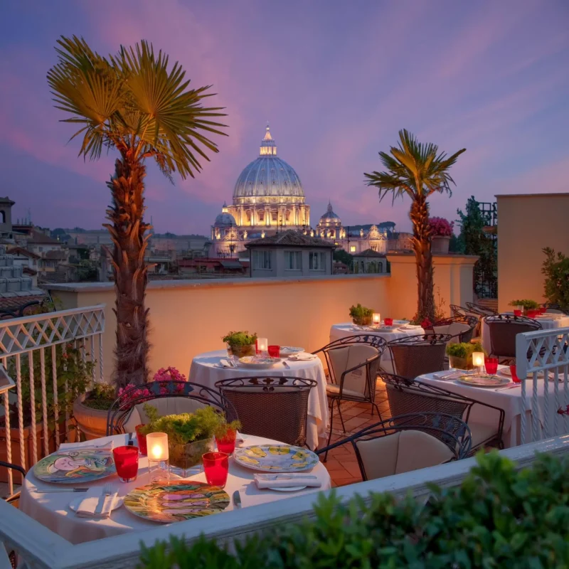 Rooftop restaurant at sunset with elegant tables adorned with candles and vibrant plates, framed by palm trees and a view of a lit-up domed building.