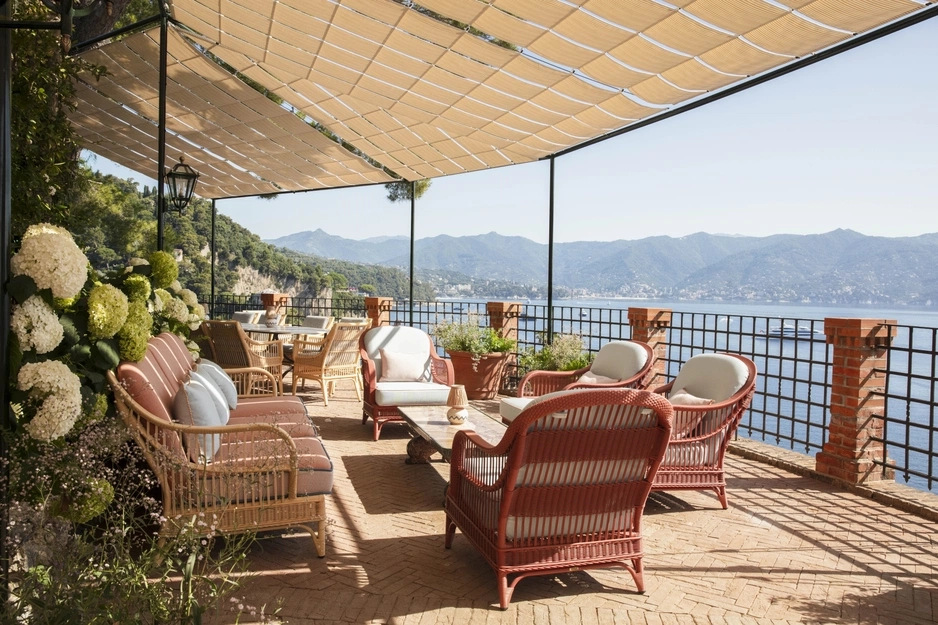 Spacious terrace with wicker seating under a shade sail, overlooking a serene ocean and distant mountains. Lush plants add a peaceful ambiance.