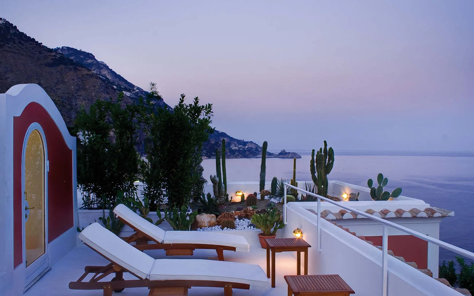 Rooftop terrace at sunset overlooking a calm sea with distant mountains. Two loungers and small tables are surrounded by cacti and soft lighting. Calm atmosphere.