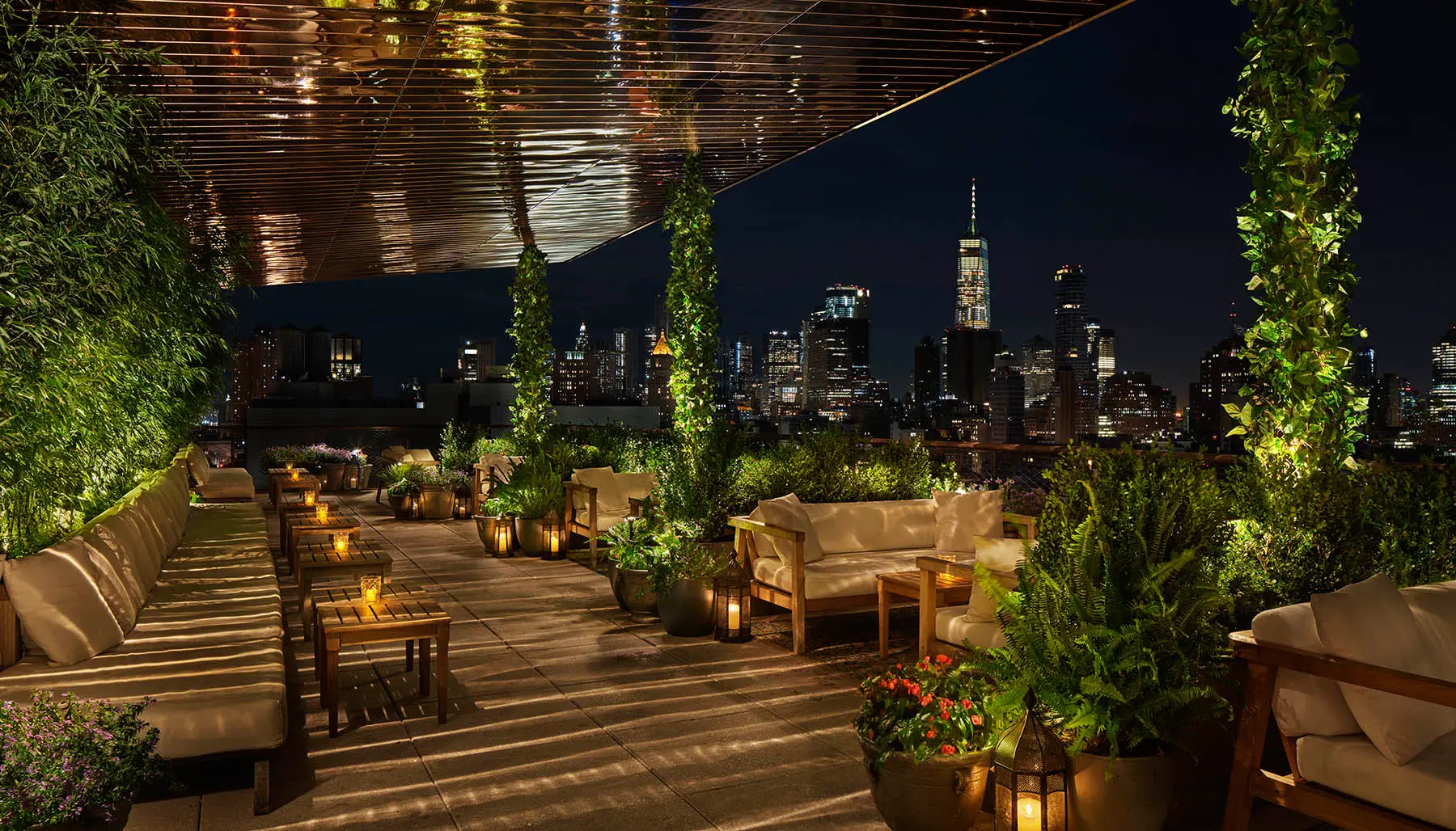 A cozy, plant-filled rooftop patio at night, with elegant seating and warm candle lights. The city skyline, crowned by a tall building, glows in the background, very common in urban rooftop escapes.