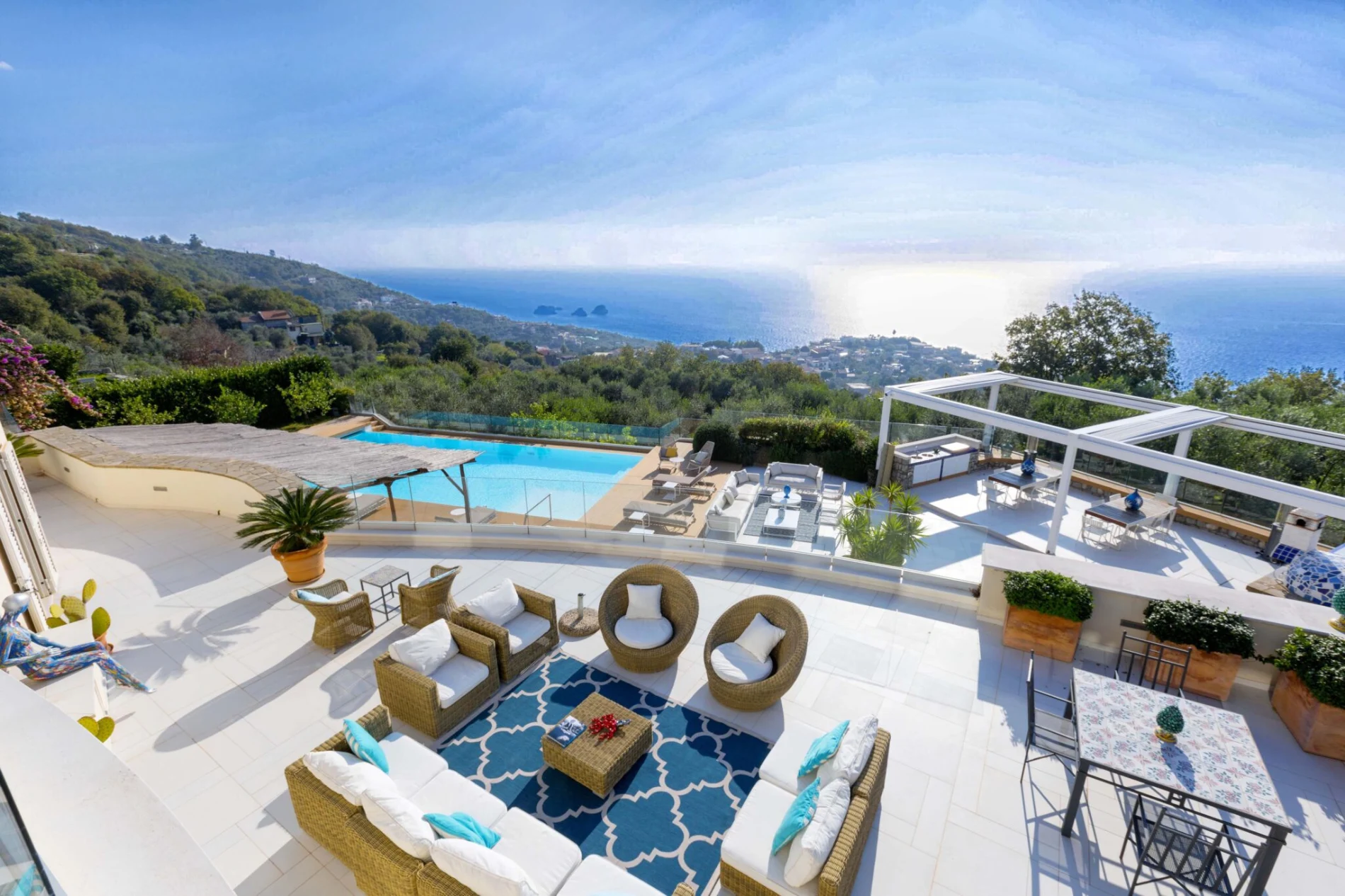 Luxurious terrace overlooking the sea, with rattan furniture, a patterned rug, pool, and shaded lounge area. Bright, serene, with a stunning ocean view.