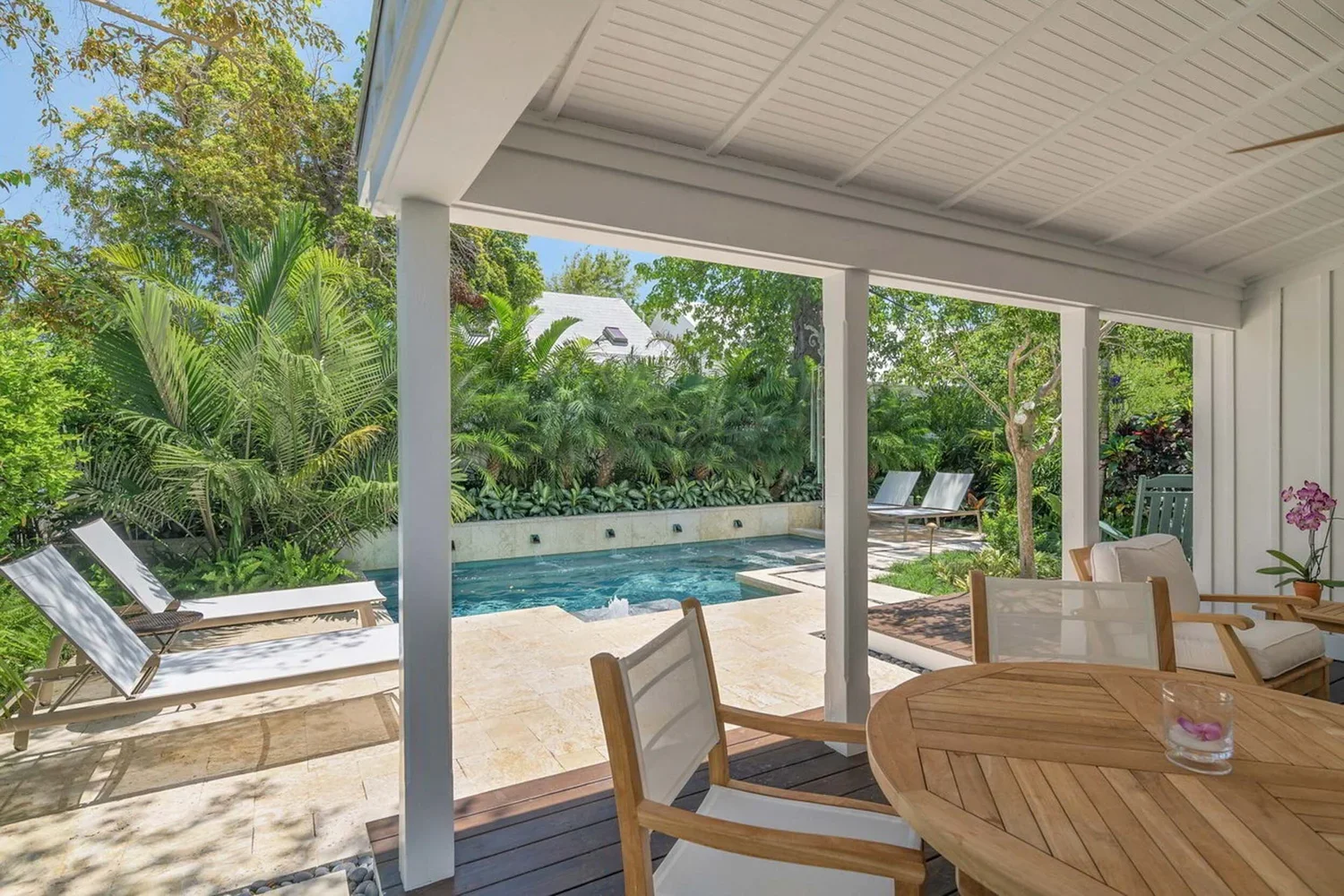 Covered patio by designer Craig Reynolds with a pool surrounded by lush greenery and palm trees, offering a view of the ocean at sunset. Relaxing and serene atmosphere. with wooden table and chairs overlooking a serene pool surrounded by lush greenery and lounge chairs, evoking a relaxing ambiance.