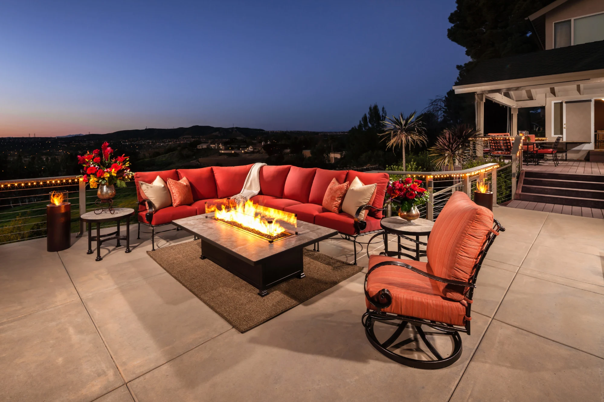 Cozy patio with red seating, a fire pit, and vibrant flowers. Overlooks a serene landscape at dusk, creating a warm and inviting atmosphere.