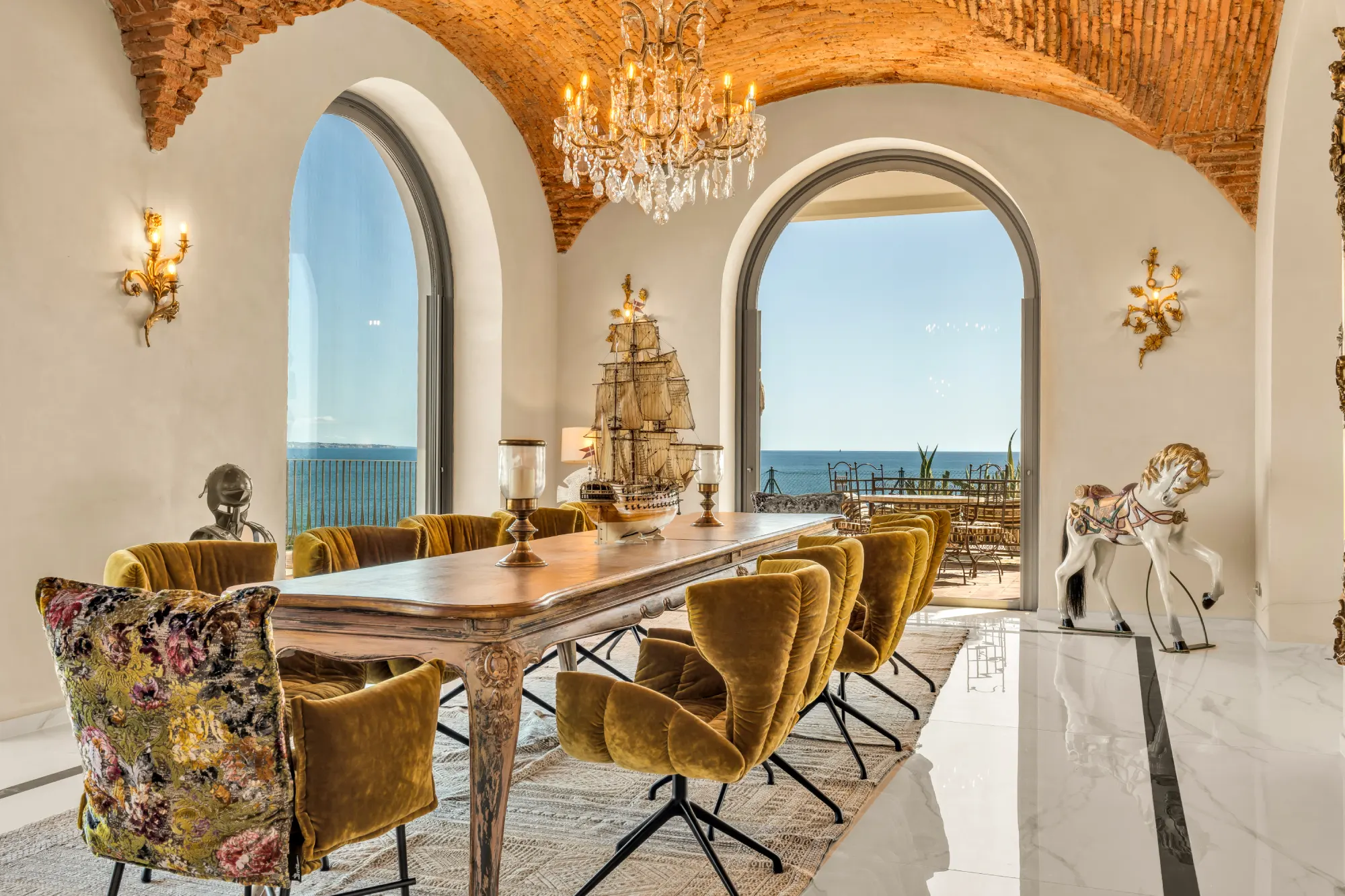 Elegant dining room with a long wooden table, plush mustard chairs, and a chandelier. Large arched windows reveal a stunning ocean view.