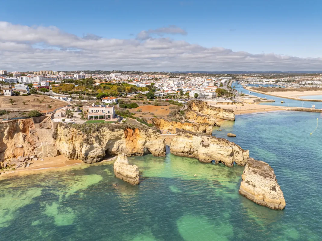 Coastal view of vibrant cliffs and clear turquoise waters in Algarve, with a small city in the background under a partly cloudy sky. The scene feels serene and inviting.