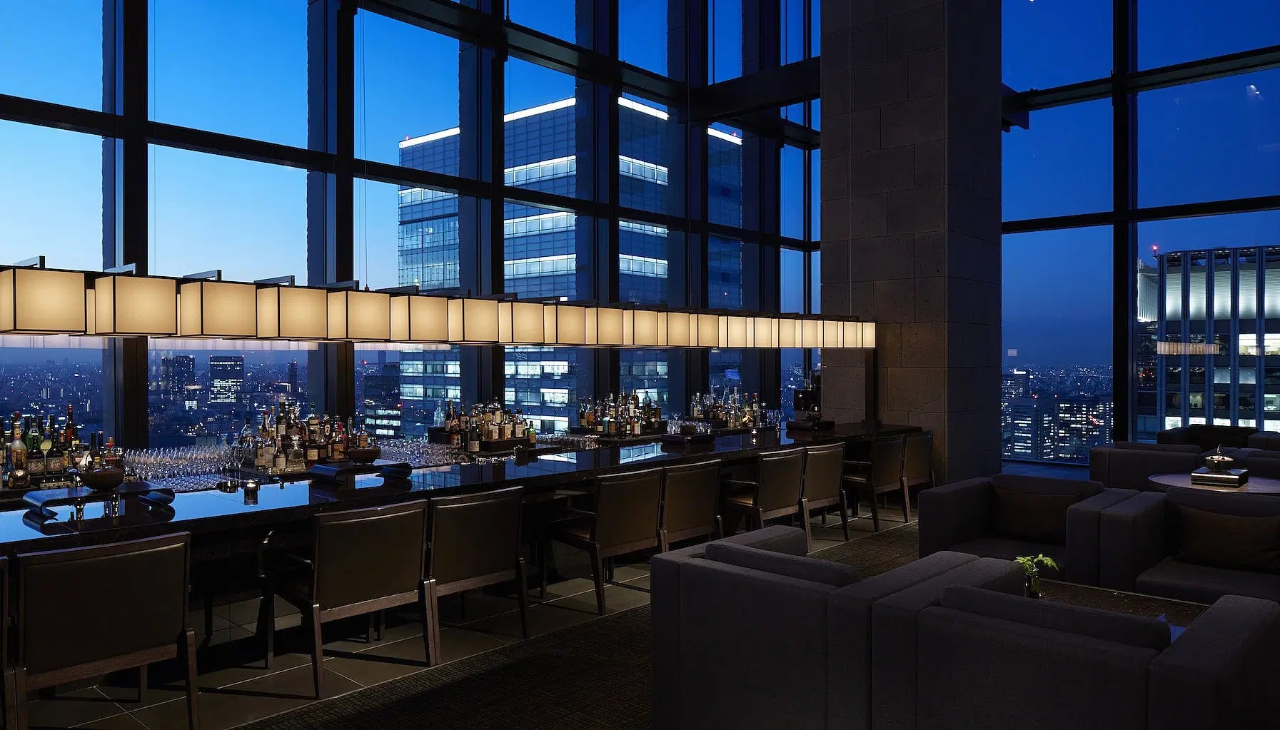Luxurious rooftop bar at dusk with city skyline view. Elegant seating, dim lighting, and reflective surfaces create a sophisticated ambiance.