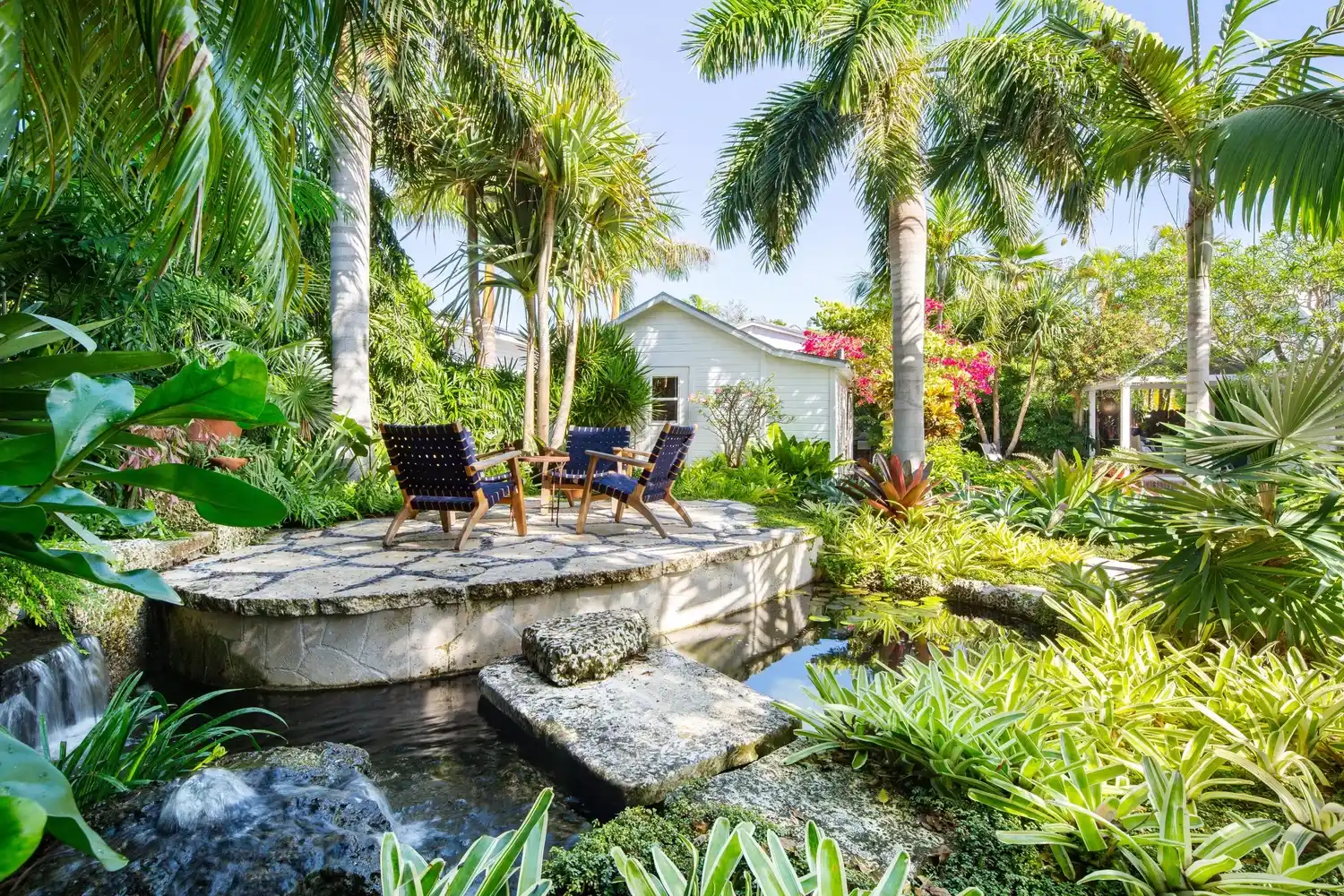 Tropical garden by designer Craig Reynolds with blue chairs on a stone patio, surrounded by lush greenery and palm trees. A serene pond flows gently beneath the patio.