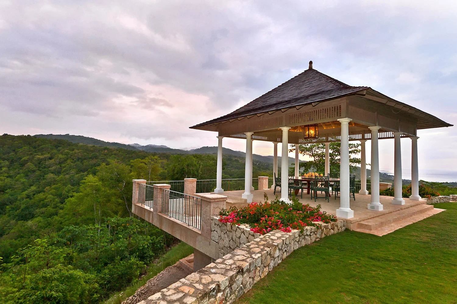 A serene open pavilion in Jamaica by designer Craig Reynolds with a wooden roof and white columns stands on a grassy hilltop, offering panoramic views of lush green forests under a cloudy sky.