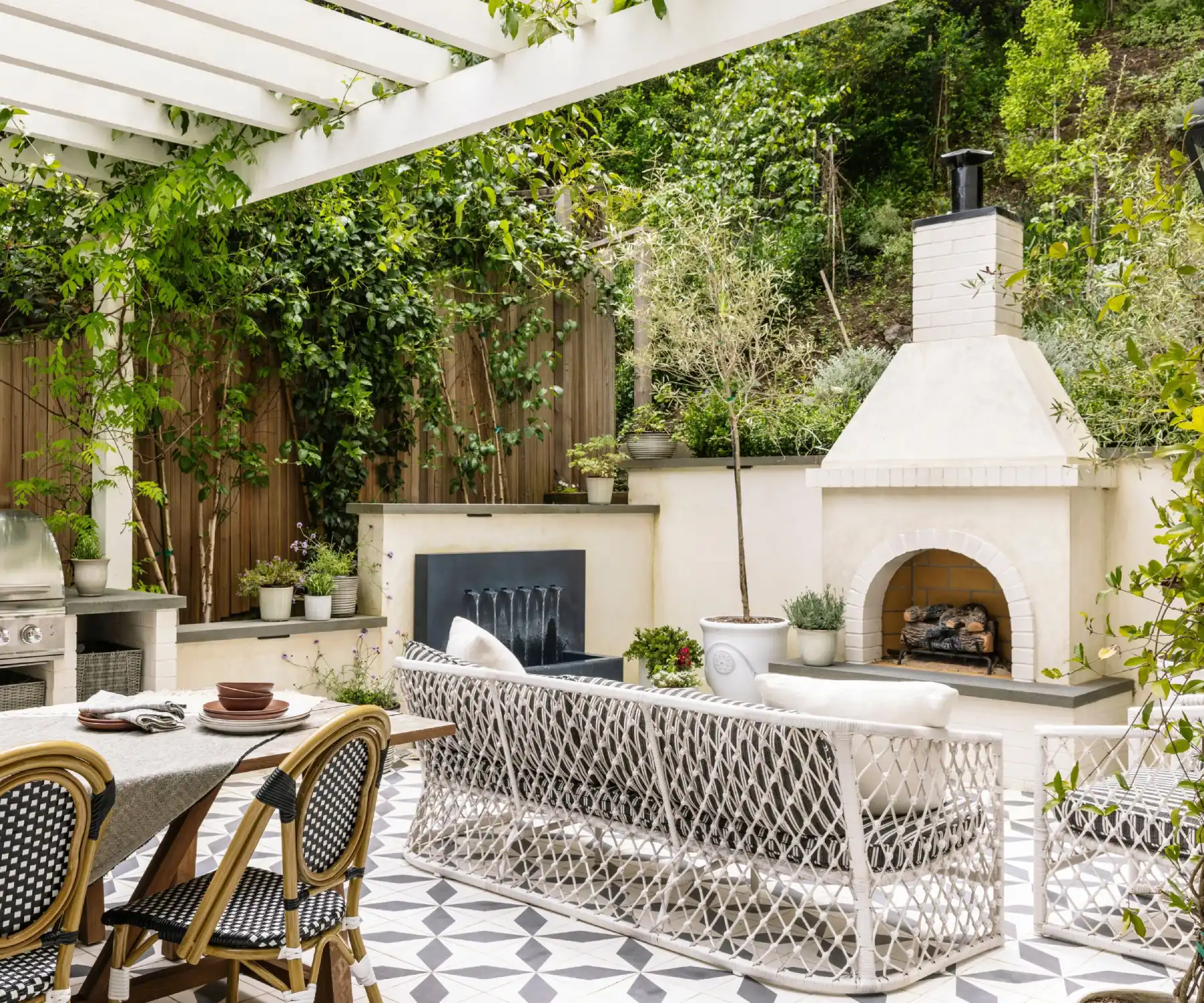 A chic outdoor patio featuring a white pergola with greenery, a cozy white fireplace, a patterned tiled floor, and stylish seating including a sofa and dining set.