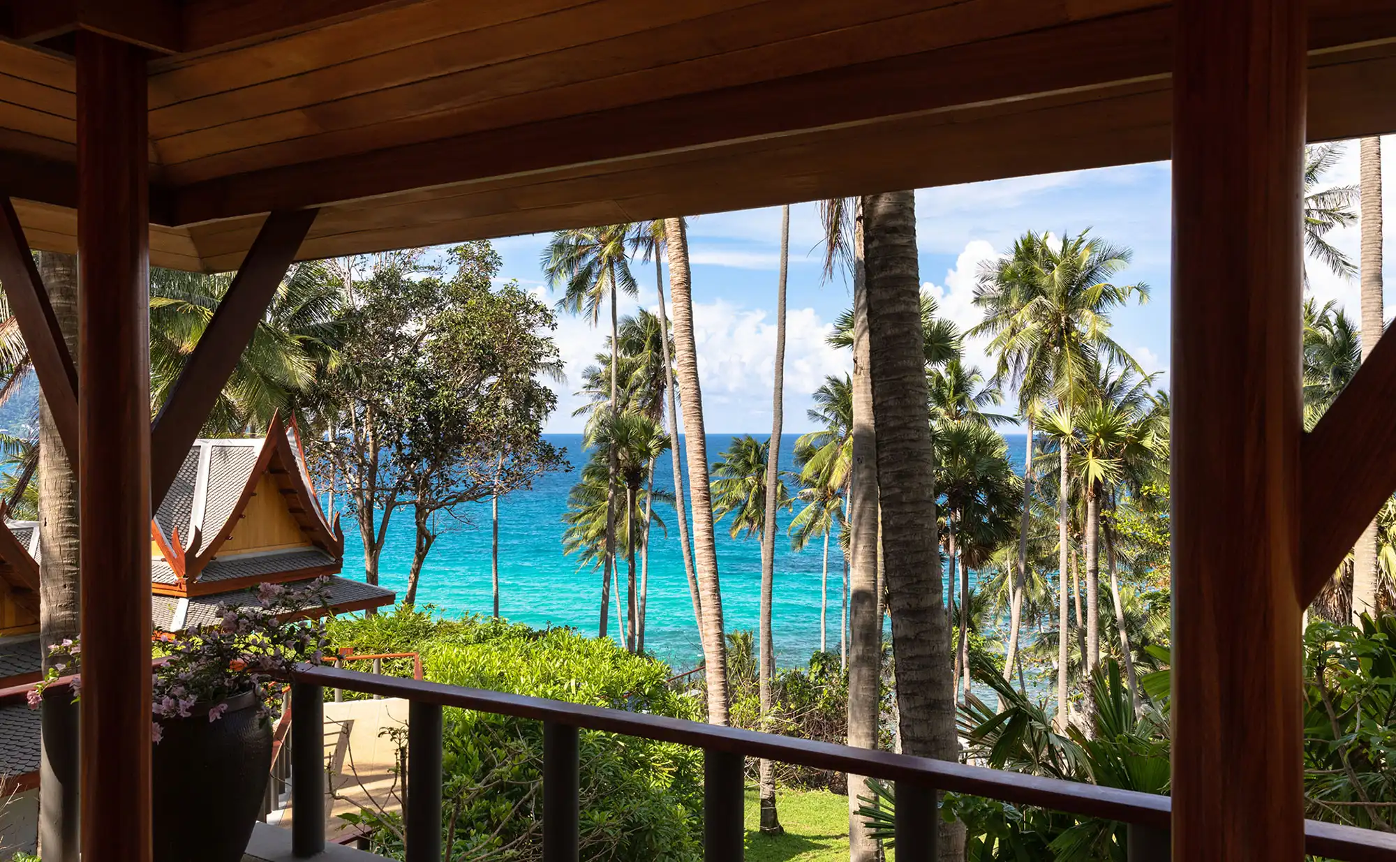 View from a wooden balcony overlooking tall palm trees, a lush garden, and a vibrant blue ocean under a clear sky, conveying a tropical paradise vibe.