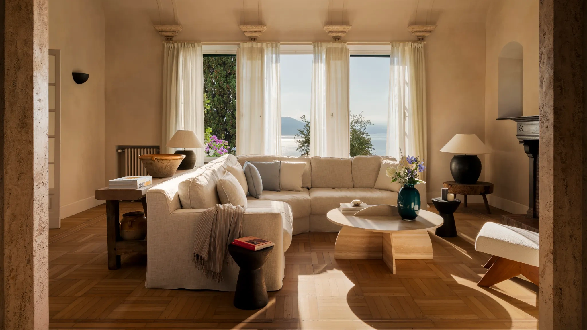 A cozy living room with a beige sectional sofa faces three large windows with sheer curtains, overlooking a serene ocean view. Warm light fills the space, enhancing the wooden floor and soft-toned walls. A round wooden coffee table, adorned with a vase of flowers, and stylish side tables complete the relaxed, elegant atmosphere.