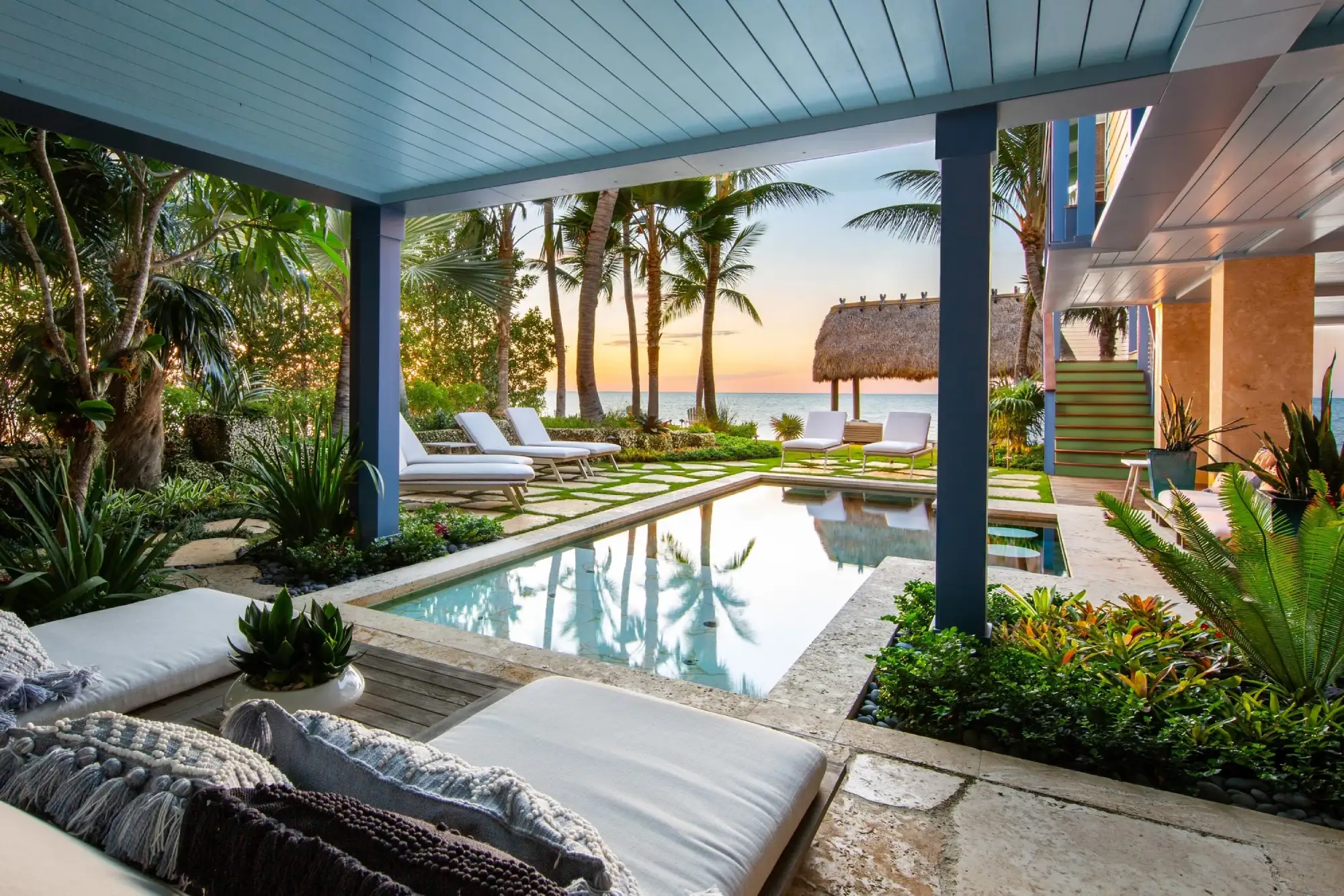 Tropical patio by designer Craig Reynolds with a pool surrounded by lush greenery and palm trees, offering a view of the ocean at sunset. Relaxing and serene atmosphere.