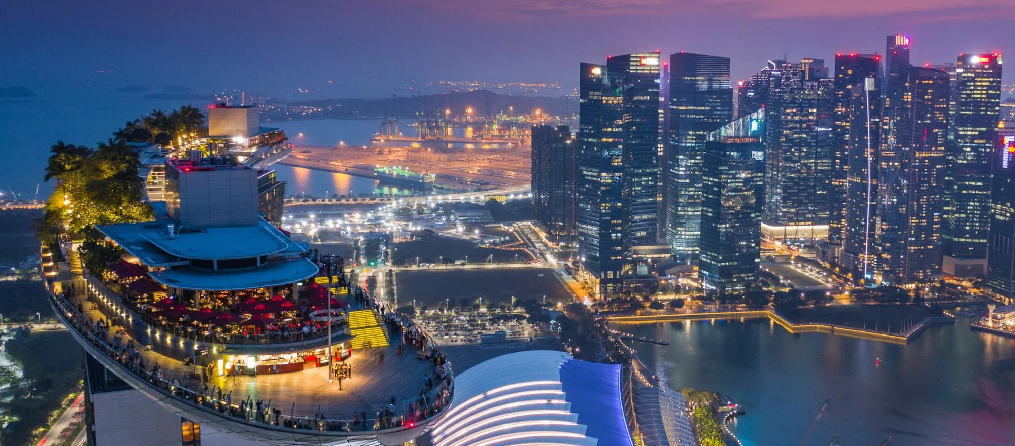 Rooftop view of a bustling cityscape at dusk, featuring modern skyscrapers with bright lights. A vibrant rooftop bar with people creates a lively atmosphere.