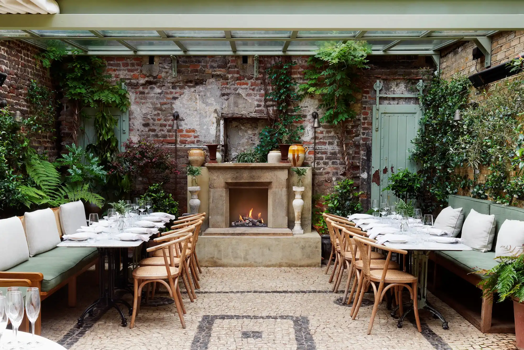 Charming courtyard with rustic brick walls, green foliage, and a lit stone fireplace. Tables set with white linens and cushioned seating. Cozy ambiance.
