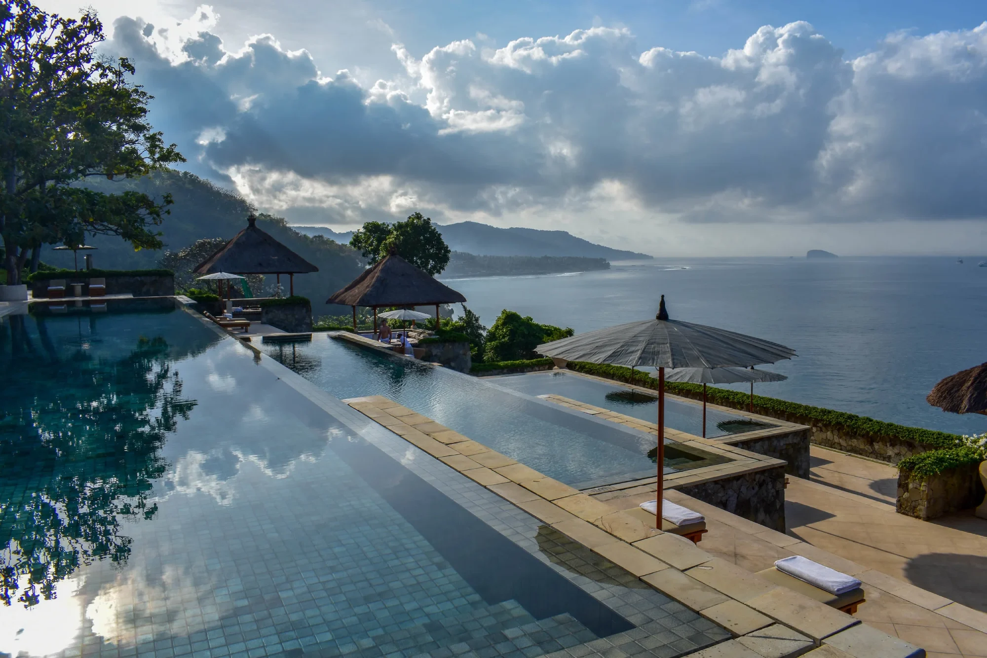 A serene infinity pool at Amankila Hotel overlooks a vast ocean, bordered by lush hills under a cloudy sky. Straw parasols and loungers add a relaxing vibe.