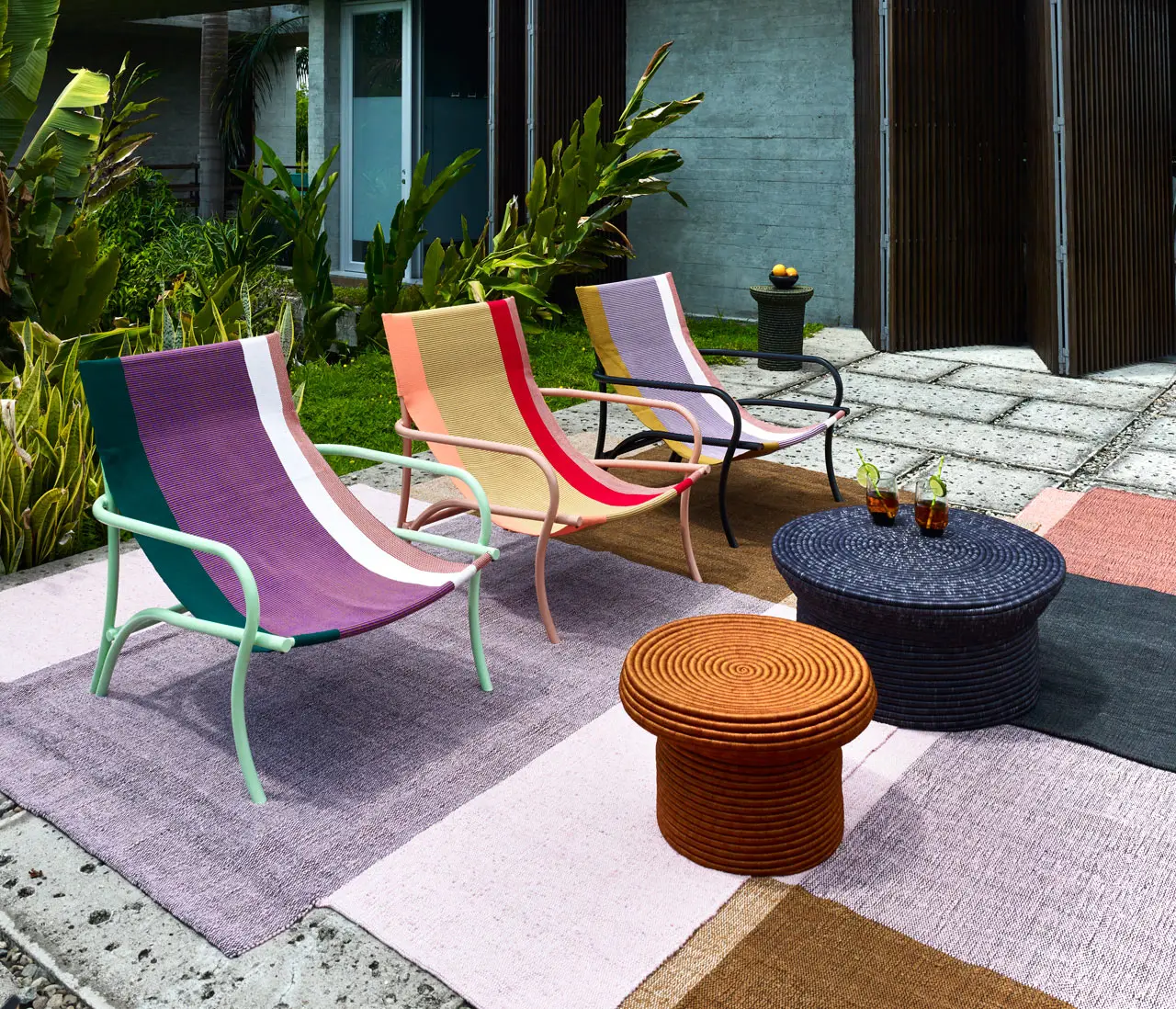 Colorful lounge chairs by Sebastian Herkner with striped patterns sit on woven rugs in a modern patio. A round table holds drinks, surrounded by lush greenery and a sleek house.
