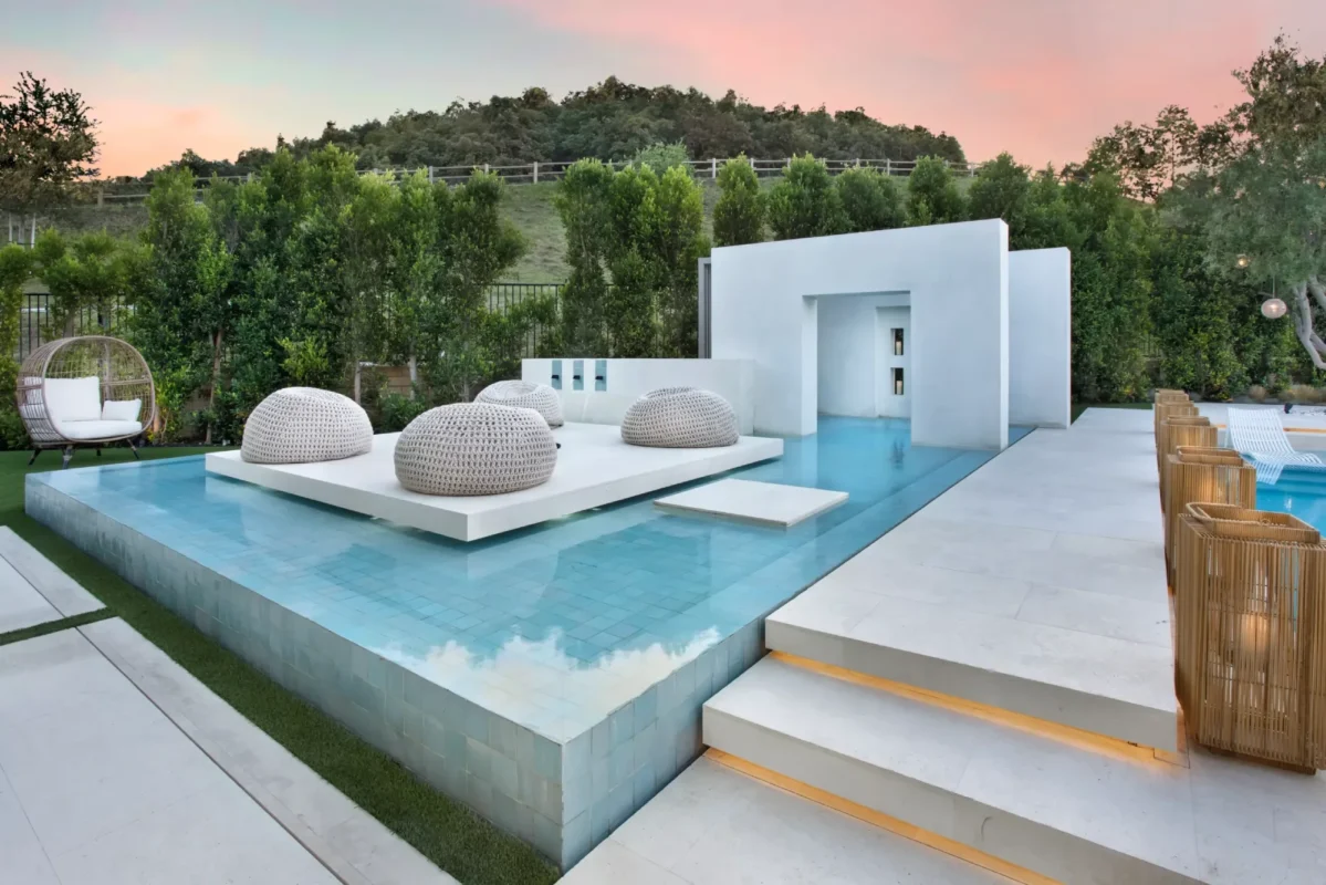 Modern outdoor lounge with a sleek design, featuring woven seats on a raised platform surrounded by a calm pool. Greenery and a vibrant sunset enhance the serene ambiance.