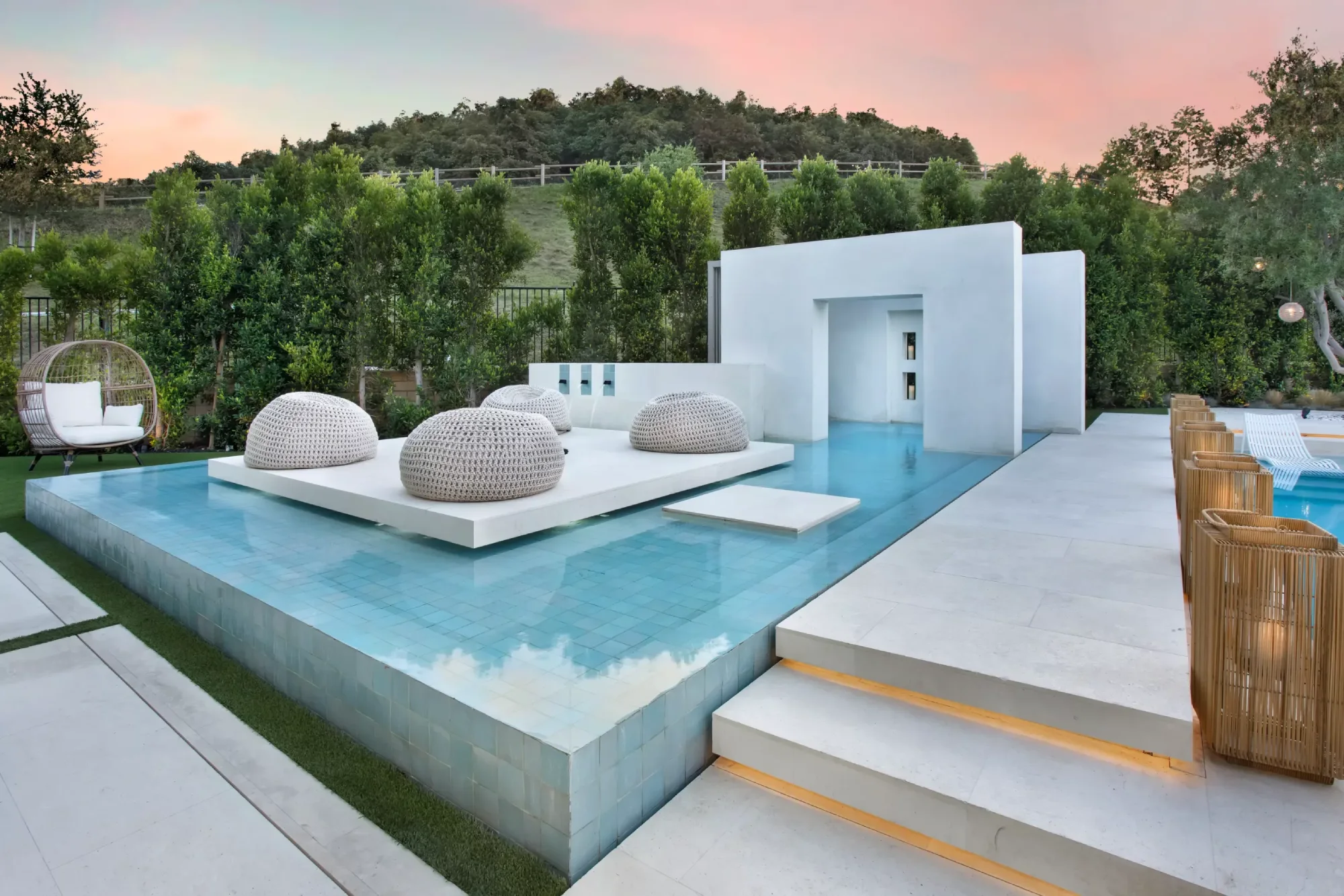 Modern outdoor lounge with a sleek design, featuring woven seats on a raised platform surrounded by a calm pool. Greenery and a vibrant sunset enhance the serene ambiance.