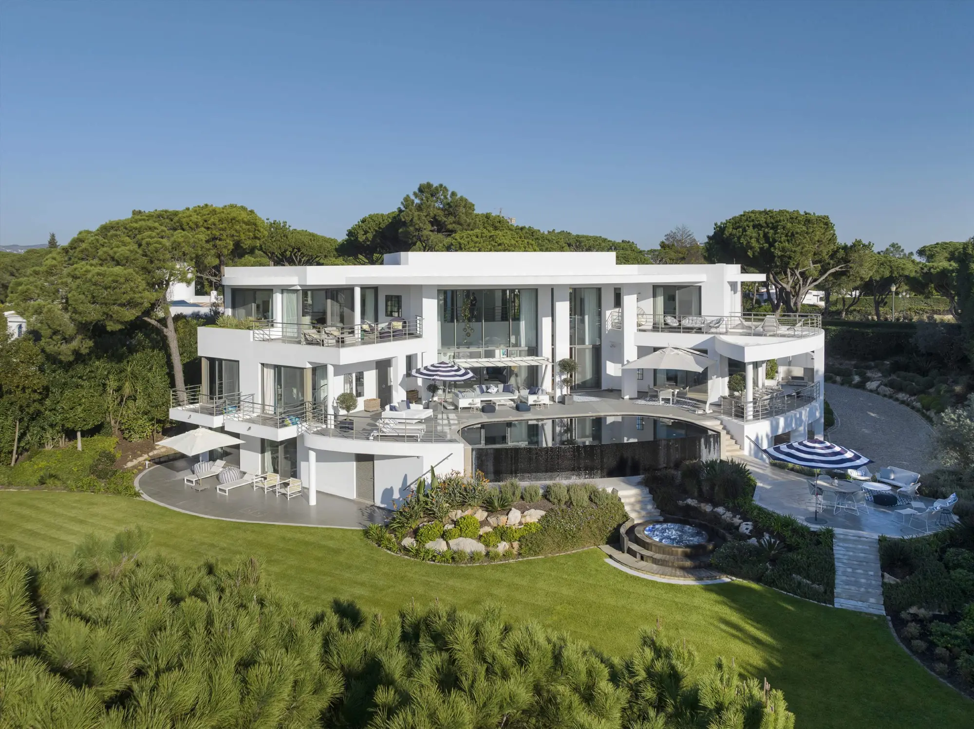 Modern white villa with large glass windows, balconies, and surrounding lush gardens under a clear blue sky, evoking luxury and tranquility, proper of Algarve luxury properties.