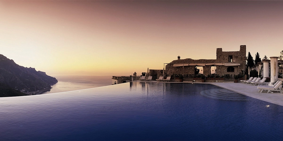 Infinity pool at sunset overlooking a vast ocean, with a rustic stone building on the right. Calm, tranquil ambiance with a gradient sky.