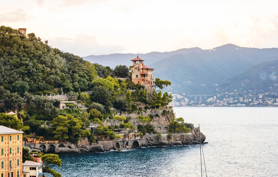 Coastal villa perched on a lush, cliffside with arched supports, overlooking a calm sea. Background features distant, hazy mountains. Serene ambiance.