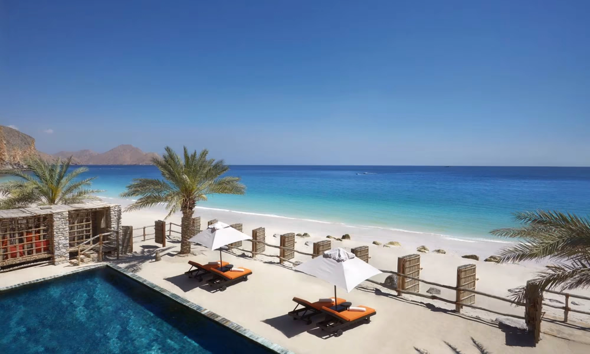 Serene beach scene with a turquoise sea, clear sky, and a sandy shore. Palm trees, sun loungers, and umbrellas line a poolside patio, conveying relaxation. This hotel is part of the top 5 seaside hotels with spectacular sea views.