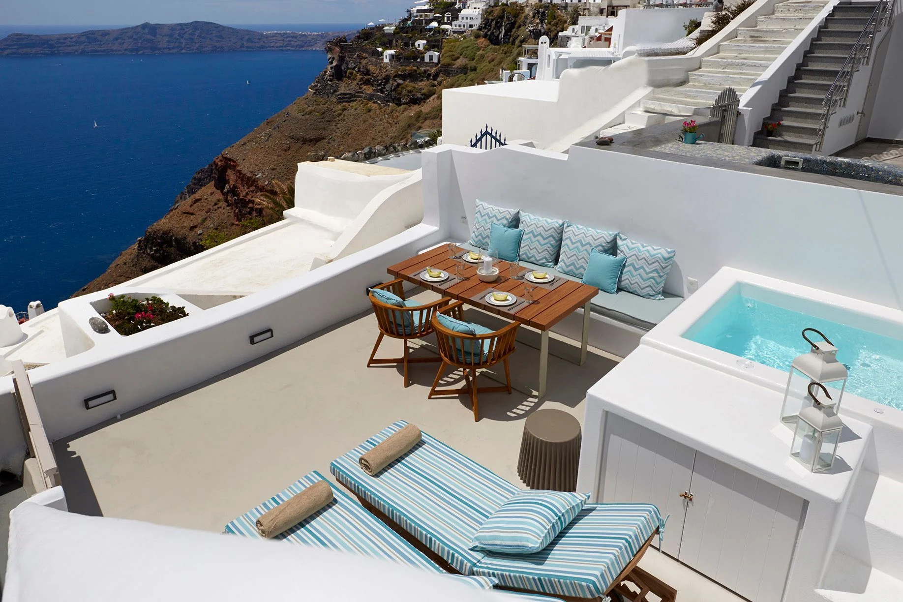 Roof terrace in Santorini with ocean view, featuring two striped loungers, a dining table with turquoise cushions, and a small pool.