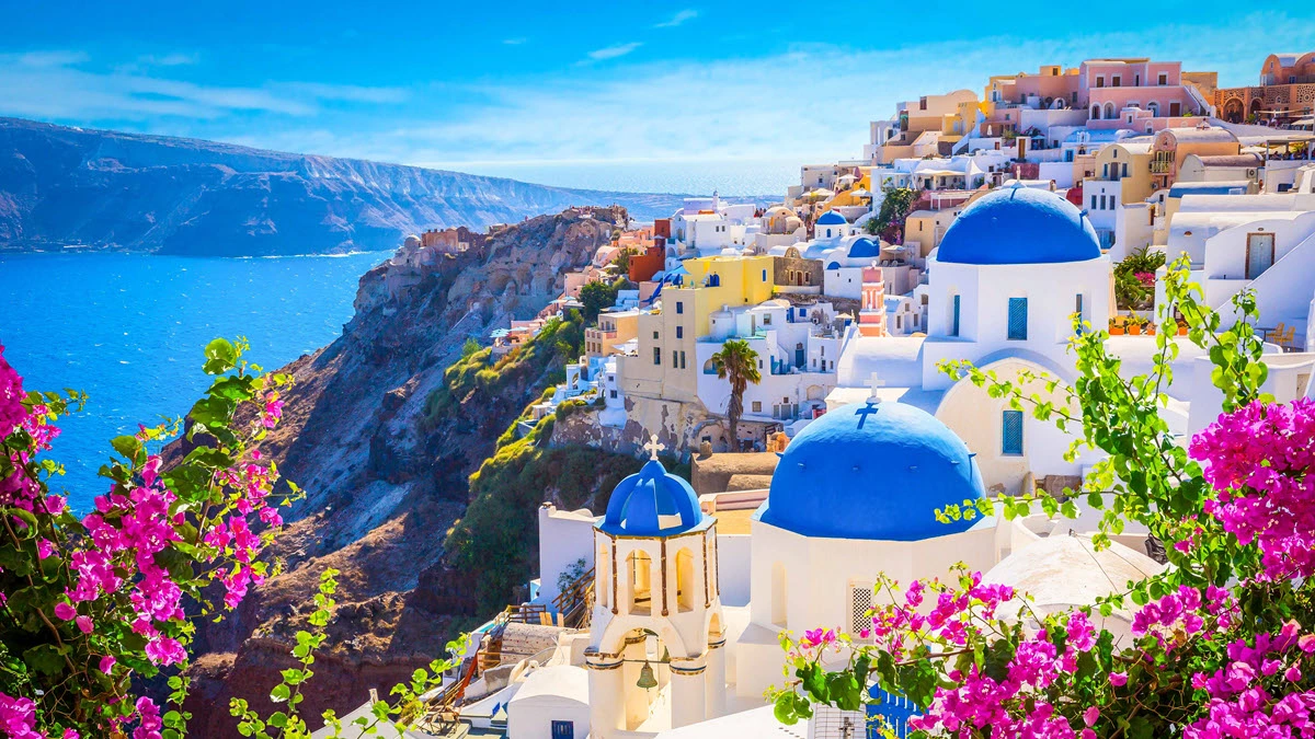 Scenic view of Santorini with whitewashed buildings and blue domes, vibrant pink flowers in the foreground, and the deep blue sea under a clear sky.