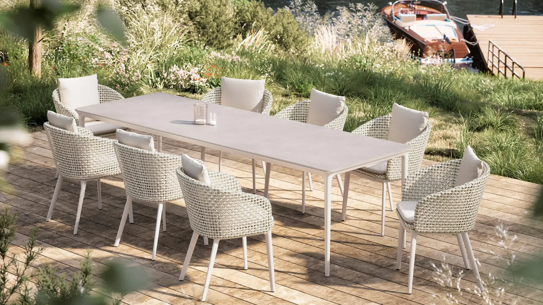 Outdoor dining setting with a long beige table and eight wicker chairs by designer Sebastian Herkner on a wooden deck. Lush greenery and a docked boat create a relaxed, summery vibe.