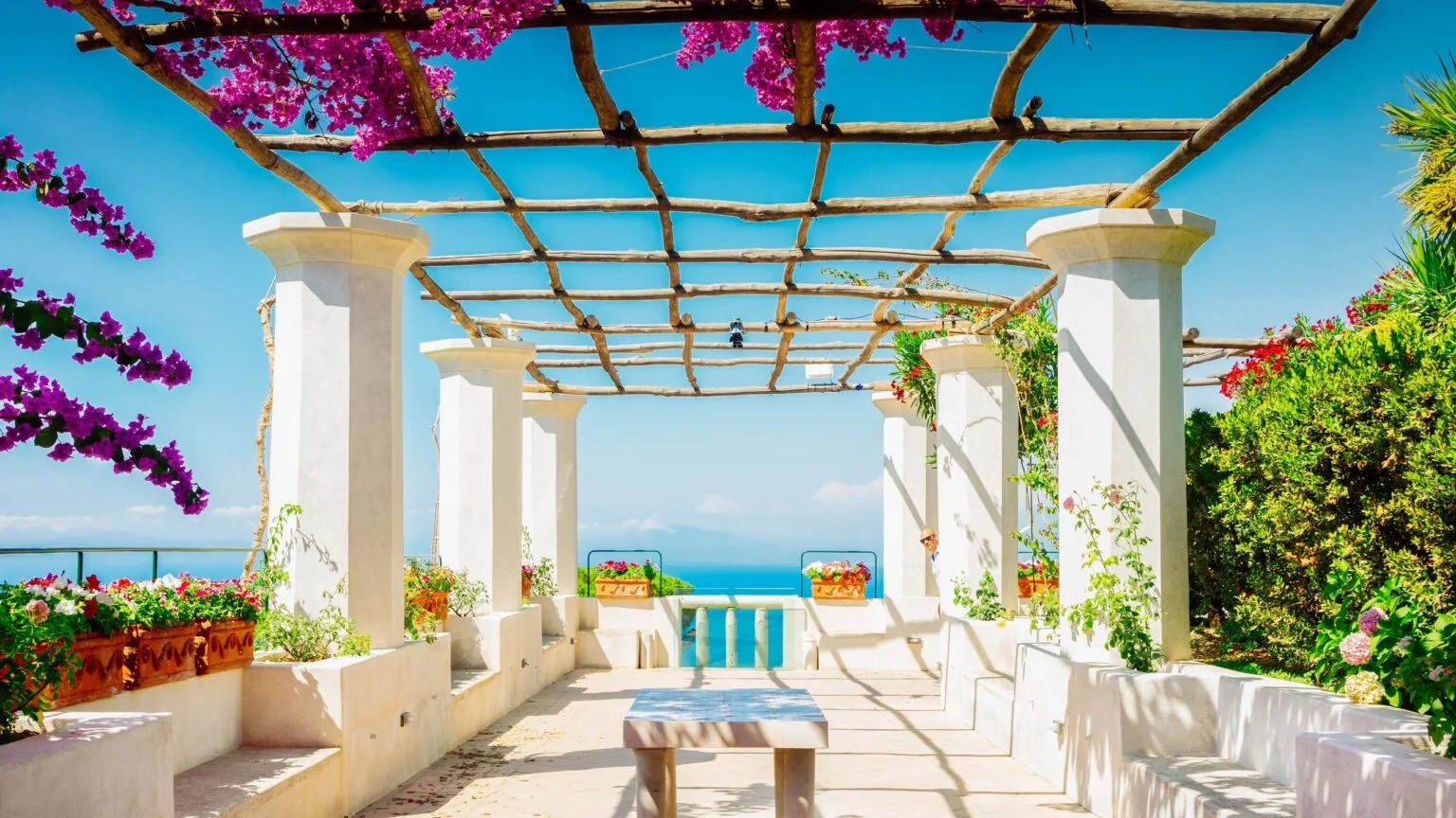 A sunny patio with white columns and a wooden pergola adorned with vibrant pink flowers, overlooking a tranquil ocean under a clear blue sky.