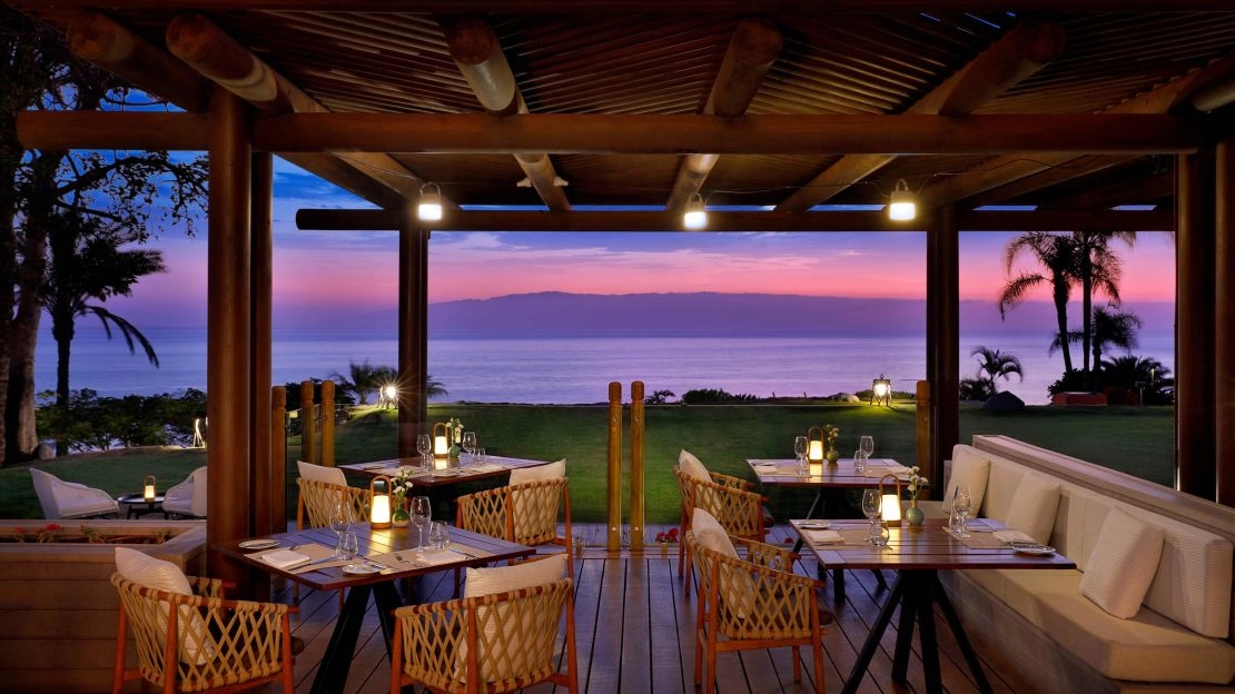 A cozy outdoor restaurant with wooden tables overlooks the ocean at sunset. The sky is vibrant pink and purple, creating a serene and romantic ambiance.