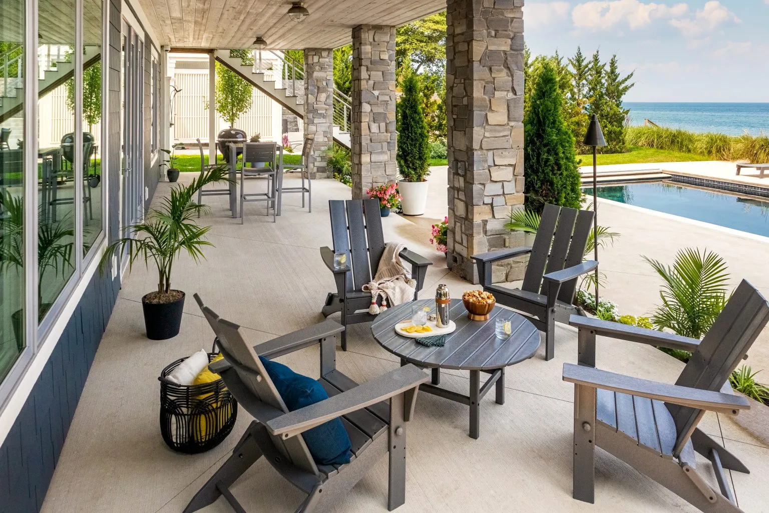 Covered patio with Adirondack chairs around a table, near a pool. Stone columns and potted plants add elegance, highlighting properly maintained outdoor furniture.