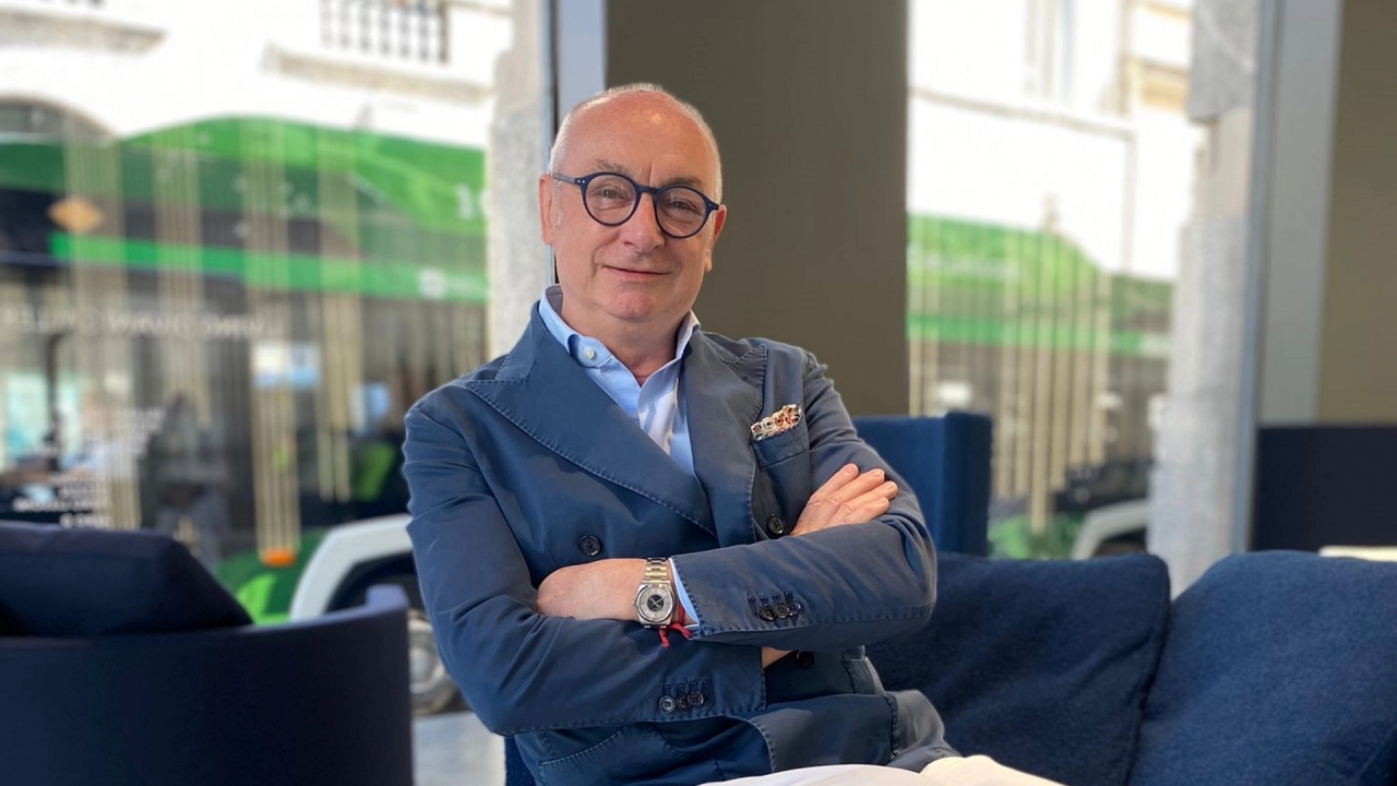 Piero Lissoni in a blue blazer and glasses sits confidently with arms crossed on a blue couch in a modern room. A green bus is visible through the large window behind him.