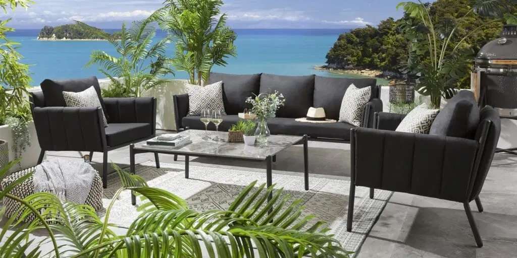 Outdoor patio with black sofas and armchairs, a glass-top coffee table, tropical plants, and ocean view. Calm and inviting atmosphere.