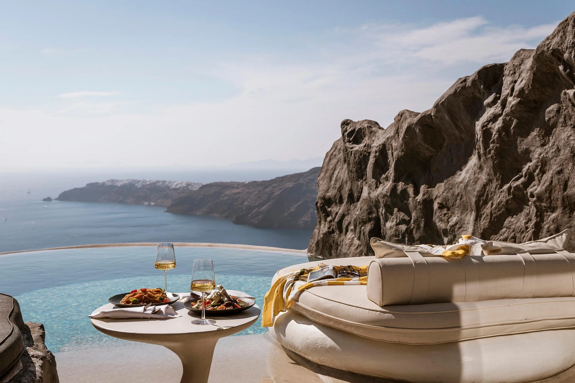 Luxurious poolside setting overlooking a serene sea, featuring a round lounge chair and a small table with two plates of food and wine glasses, highlighting santorini’s luxury cliff residences.